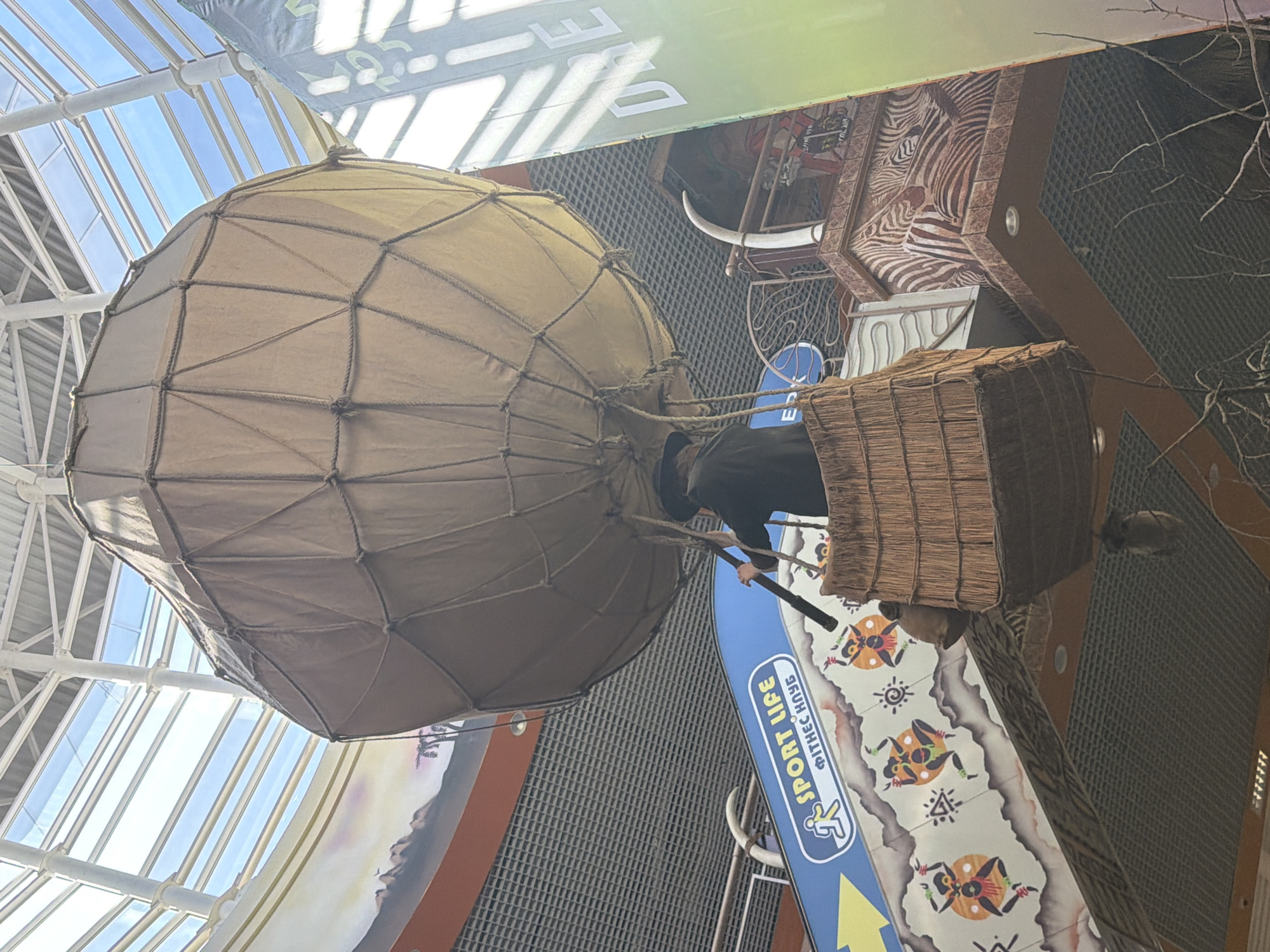 The image shows a decorative hot air balloon suspended indoors, likely in a shopping mall or entertainment complex. The balloon is made of tan fabric with a rope netting and is suspended above a basket-like gondola. A person wearing a dark hat and coat is standing in the gondola, holding a long pole. Below the balloon, a blue banner with yellow lettering that reads "SPORT LIFE" and "ФІТНЕС КЛУБ" (Fitness Club) is visible, along with a stylized depiction of people dancing. Further down, there are decorative elements resembling tribal art, a zebra-patterned wall, and what appears to be tusks. The ceiling structure of the building, with white beams and a glass roof, is visible at the top of the image. The overall setting suggests an indoor recreational or commercial space. The "SPORT LIFE" banner indicates this is likely within one of their fitness club locations in Kyiv, Ukraine.