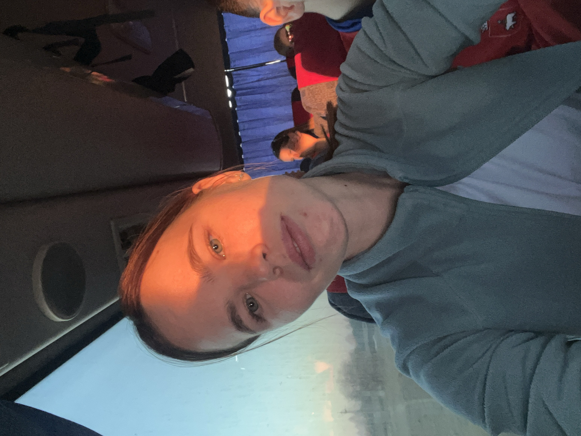 A selfie photograph taken inside a vehicle. The main subject is a person with fair skin and light-colored eyes, wearing a gray-blue long-sleeved shirt over a white t-shirt. The person's face is illuminated by sunlight, creating shadows. Other people are visible in the background, sitting behind blue window coverings. The vehicle appears to be in motion, as suggested by the blurry view through the window. The location, Ustinovo, Russia, cannot be confirmed.