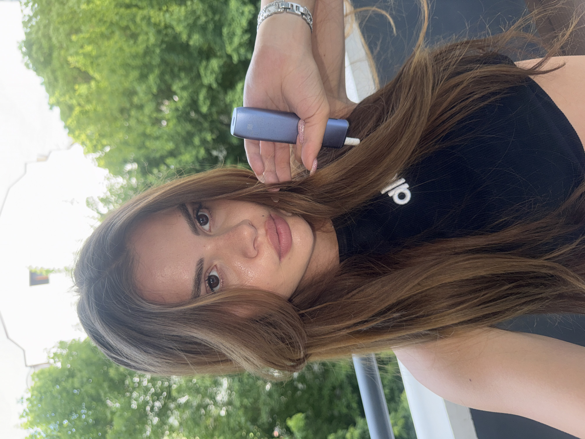 A medium close-up, rotated image shows a young woman with long, wavy, medium-brown hair looking directly at the camera. Her head is tilted, and her expression is neutral, with full lips and dark eyes. Her skin appears dewy and lightly made up.
She is wearing a black t-shirt, with the white "adidas" logo partially visible on her right chest area. A silver-toned bracelet with clear, sparkling stones adorns her left wrist. Her fingernails are painted with glittery pink nail polish.
In her right hand, she holds a small, rectangular, bluish-gray electronic device. A white, cigarette-like stick protrudes from one end of the device, suggesting it might be a heated tobacco product or a vape. Her hand is positioned near her hair, framing the device prominently.
The background is softly blurred, displaying vibrant green foliage from trees, indicating an outdoor setting. The bright light suggests it is daytime, possibly with an overcast sky or dappled sunlight creating bokeh effects in the background. The overall image gives the impression of a casual outdoor portrait or selfie.