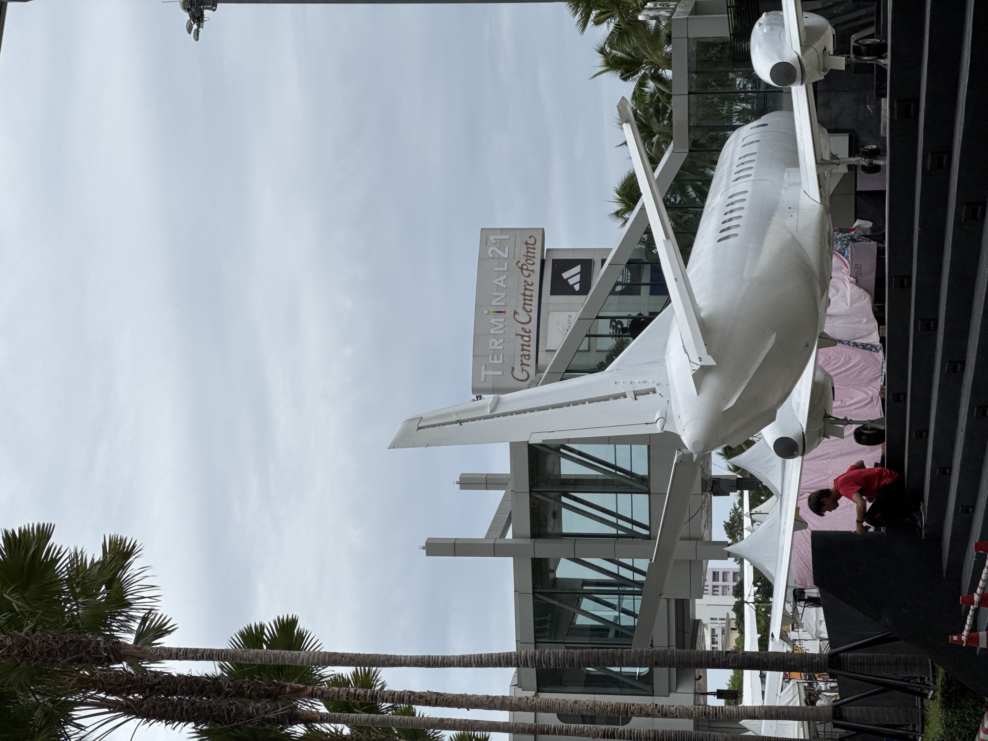 In Pattaya City, Thailand, an outdoor scene features a white airplane model serving as a prominent display. The aircraft is positioned on a dark, tiered platform with stairs leading up to it, in front of a modern building complex. A pedestrian walkway, enclosed in glass and steel, is visible connecting to the building. Tall palm trees line the left side of the frame, adding a tropical element to the urban setting. The sky is overcast and gray.
A single individual, wearing a red shirt and dark pants, is seated on the steps of the platform, facing the airplane and appearing to be engaged with something in their lap. Behind the airplane and the person, white tent-like structures are set up, suggesting a market or event. The building complex behind the airplane has a large sign that reads "TERMINAL 21 Grande Centre Point" along with an Adidas logo. Further architectural details of the building, including large glass windows and modern support structures, are visible. The overall atmosphere is one of an urban public space with an artistic or commercial installation.