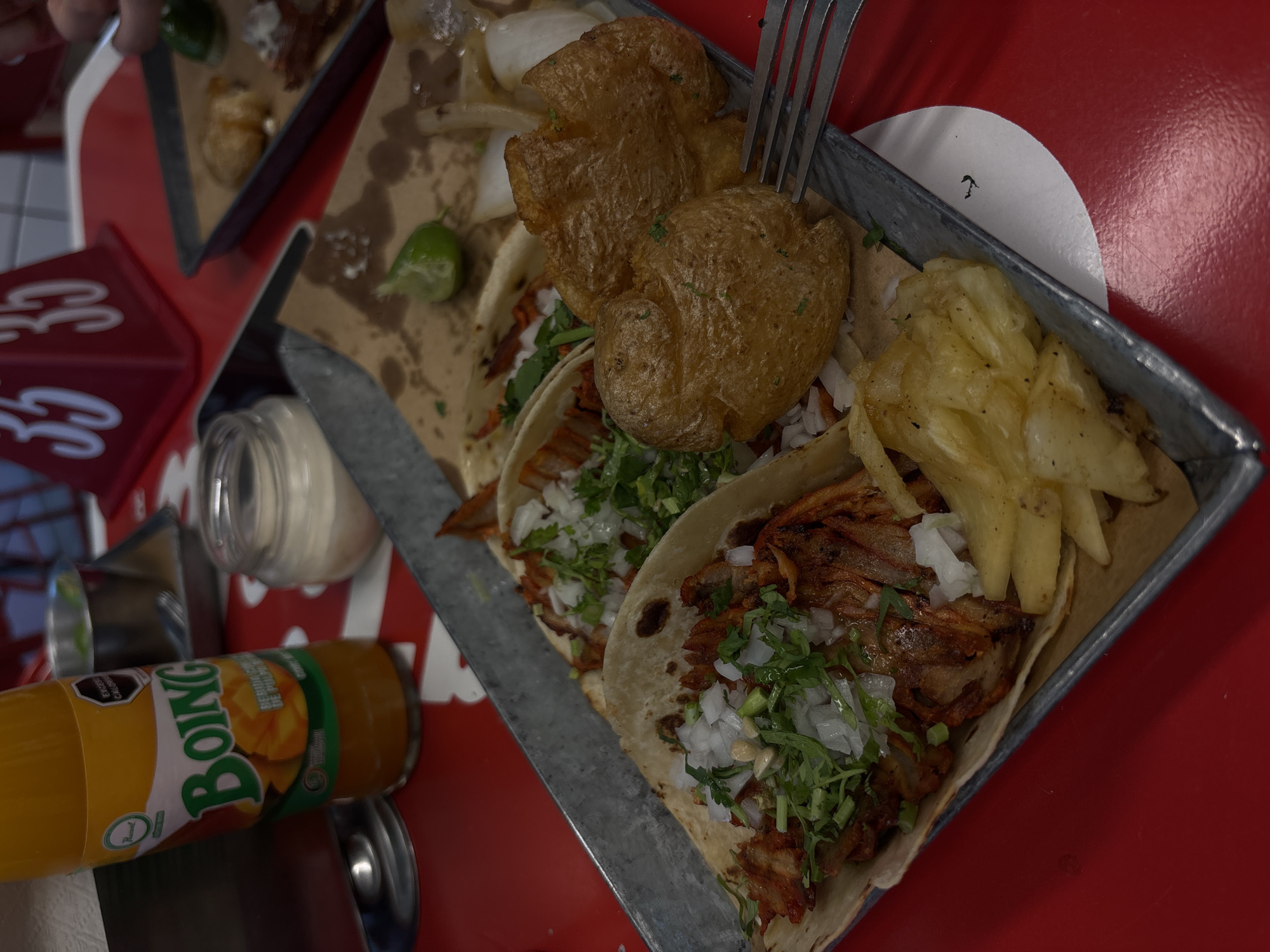 The image depicts a close-up, high-angle view of a metal serving tray filled with tacos, fries, and potatoes. The tacos are topped with chopped onions and cilantro. A fork is seen positioned near the food. To the left, a glass jar and a bottle of Boing mango drink are visible on a red table. Based on the type of food and drink present, as well as the name of the drink (Boing), it's highly likely this image was taken in Mexico City, Mexico. The scene suggests a casual dining environment.