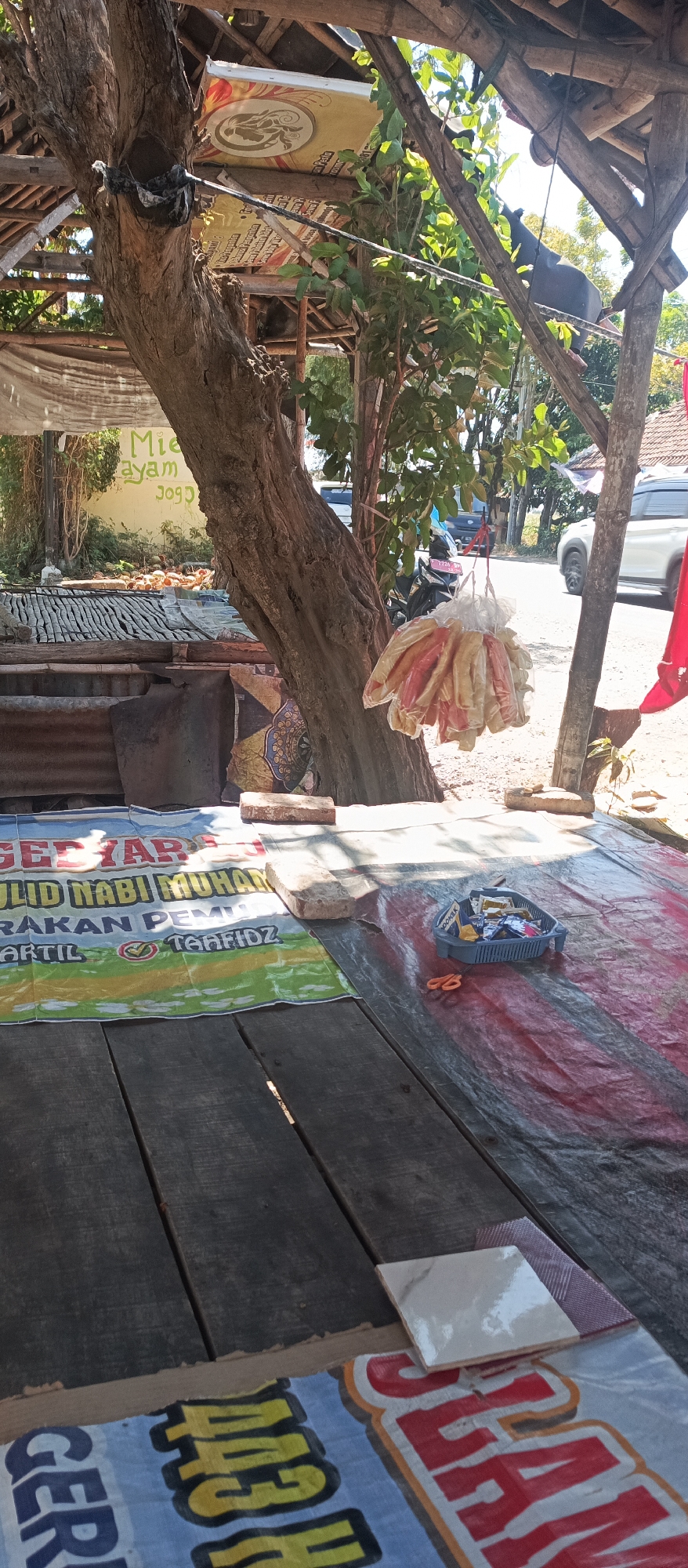 The image shows a street vendor stall in Jatisari, Indonesia. In the foreground, a wooden table is covered with a red and black tarp. On the tarp, there is a blue plastic basket containing various items, including small packets and what appear to be snacks. A pair of orange scissors is also visible next to the basket. To the left of the table, a banner with Indonesian text is laid out. The background features a large tree, a small corrugated metal structure, and foliage. A street with vehicles, including a white car and a motorcycle, is visible in the distance, suggesting an urban or semi-urban environment. The overall scene depicts a typical roadside stall operating during daylight hours.