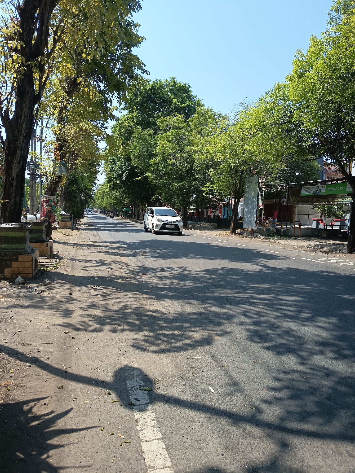 The image depicts a street scene in Kandangjati Kulon, Indonesia, on a clear, sunny day. A white sedan is traveling on the right side of the asphalt road, which is lined with trees casting dappled shadows across the pavement. The trees are lush and green, indicating a tropical or subtropical environment. On the left side of the road, there are some stone structures and a poster displaying text and images, possibly advertising. Further down the road, several buildings and more trees are visible, with a few motorcycles also in motion. The sky is a bright, cloudless blue. The overall impression is one of a typical urban or semi-urban street in Indonesia during daylight hours.