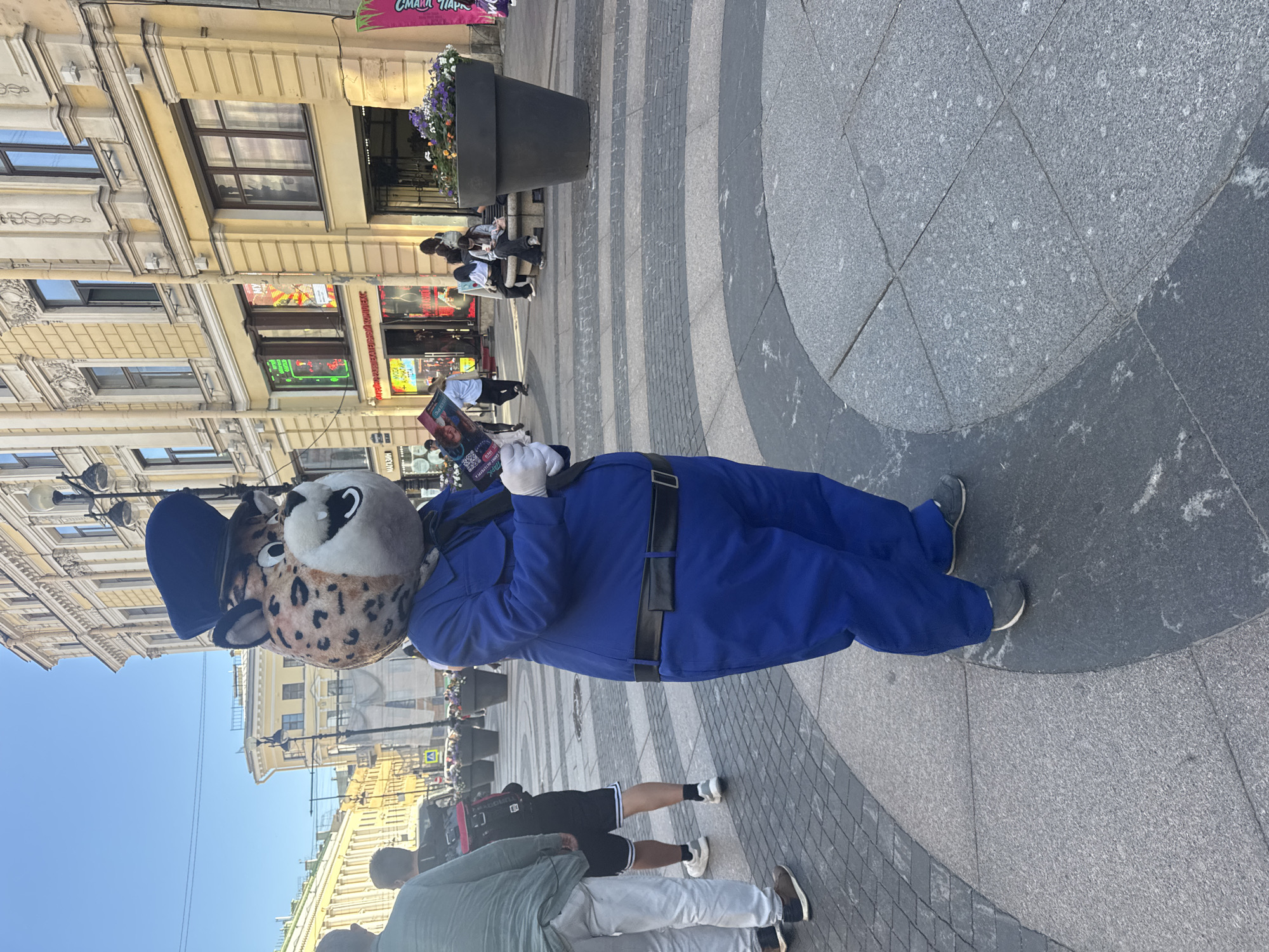 In Saint Petersburg, Russia, on Nevsky Prospekt, a person in a jaguar mascot costume stands on a wide pedestrian walkway paved with granite tiles. The mascot, wearing a blue uniform with a black belt and cap, holds leaflets in gloved hands. The background features a bright blue sky and buildings with classical architecture, characteristic of the city. Other pedestrians are visible in the distance, some sitting on benches near large planters, while others walk along the street. The overall scene depicts a lively urban environment on a sunny day.