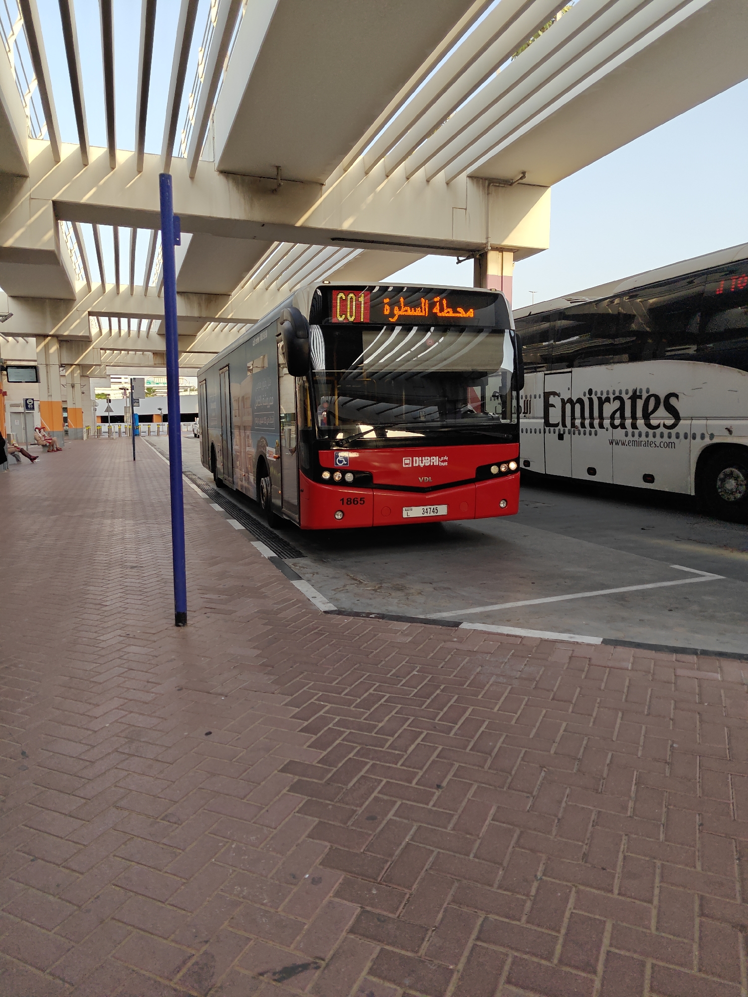 This media file captures a street scene in Dubai, United Arab Emirates, likely at a bus station. A bright red Dubai bus with "C01" displayed on its digital sign is prominently featured, suggesting it's about to depart or has just arrived. The bus also displays "محطة السطوة" in Arabic, indicating it's heading to Satwa Station.
To the right of the bus is a white Emirates coach, identifiable by the prominent "Emirates" logo and "www.emirates.com" text. This suggests the scene is at a location where both public buses and coaches operate, possibly a central transportation hub.
The foreground is paved with reddish-brown interlocking bricks laid in a herringbone pattern. A tall, slender blue pole stands vertically in the mid-ground, partially obscuring the bus and the overhead structure. The background reveals a modern, covered structure with numerous parallel beams, typical of an outdoor public space designed to offer shade. The sky is a clear, bright blue, indicating a sunny day, likely during daylight hours.
In the far background, a few people can be seen sitting on benches, waiting. The overall impression is one of a functional and bustling public transportation area in a warm climate. The presence of both a Dubai bus and an Emirates coach highlights the diverse transportation options available in the region.