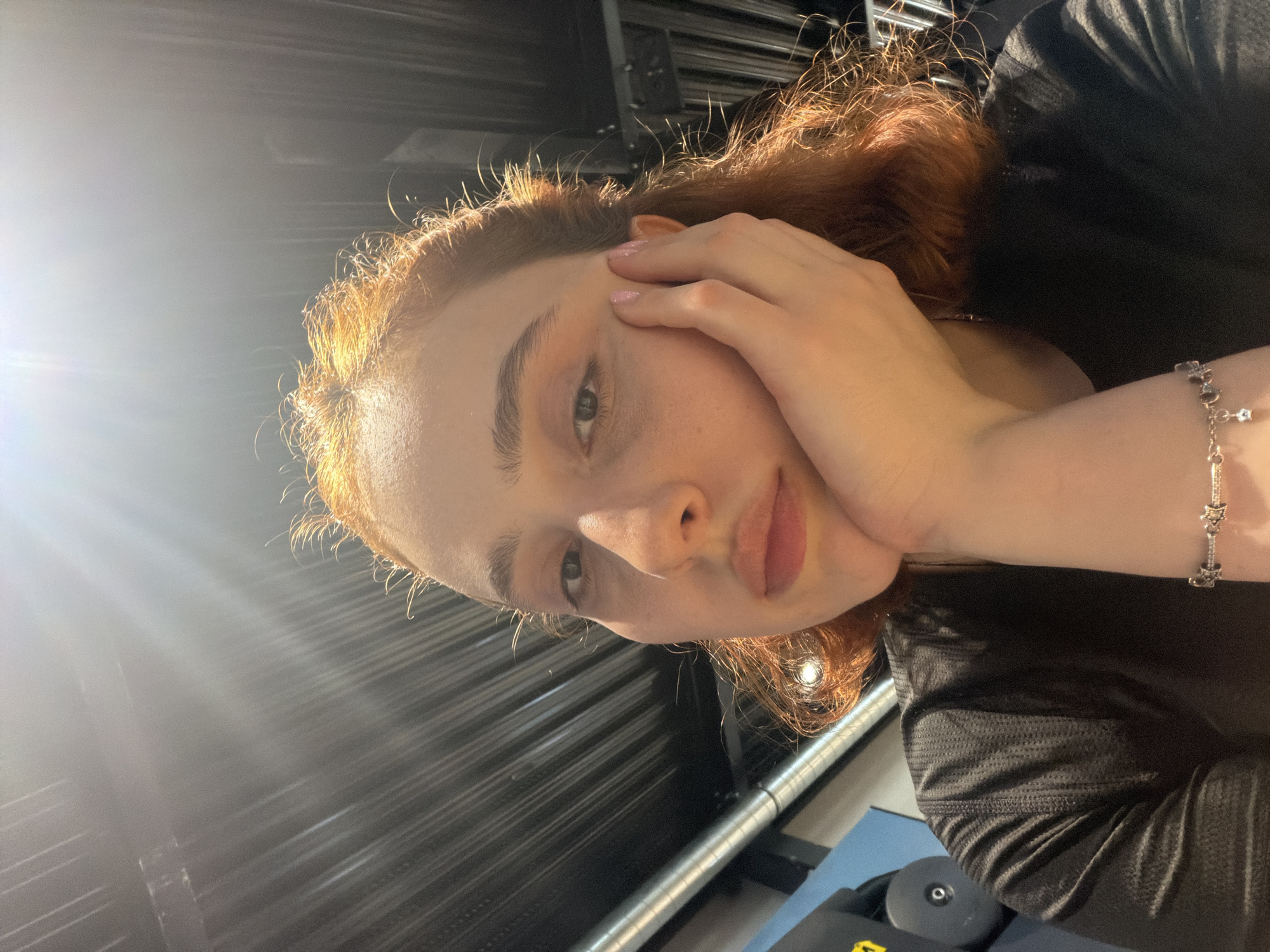 The image displays a rotated portrait of a young individual in what appears to be an indoor setting in Strasbourg, France. The subject has fair skin and prominent reddish-brown hair, which is intensely highlighted by a strong light source originating from the upper left, casting a noticeable lens flare across the frame.
The individual looks directly at the camera with a neutral, calm expression, their lips slightly parted. One hand, with light pink nail polish, is gently placed against the side of their face. A silver-colored bracelet adorned with star-shaped charms is visible on their wrist.
The person is wearing dark, likely black, clothing with a textured pattern, suggesting activewear or athletic apparel. The background features dark, horizontally ribbed or slatted panels, creating a modern or industrial aesthetic. In the bottom right corner, elements consistent with gym equipment are visible, including a dark circular component, a blue surface, and a metallic bar, strongly indicating a gym as the specific location. The pronounced backlighting creates a dramatic effect, outlining the subject and emphasizing the texture of their hair. No visible text is present in the image.