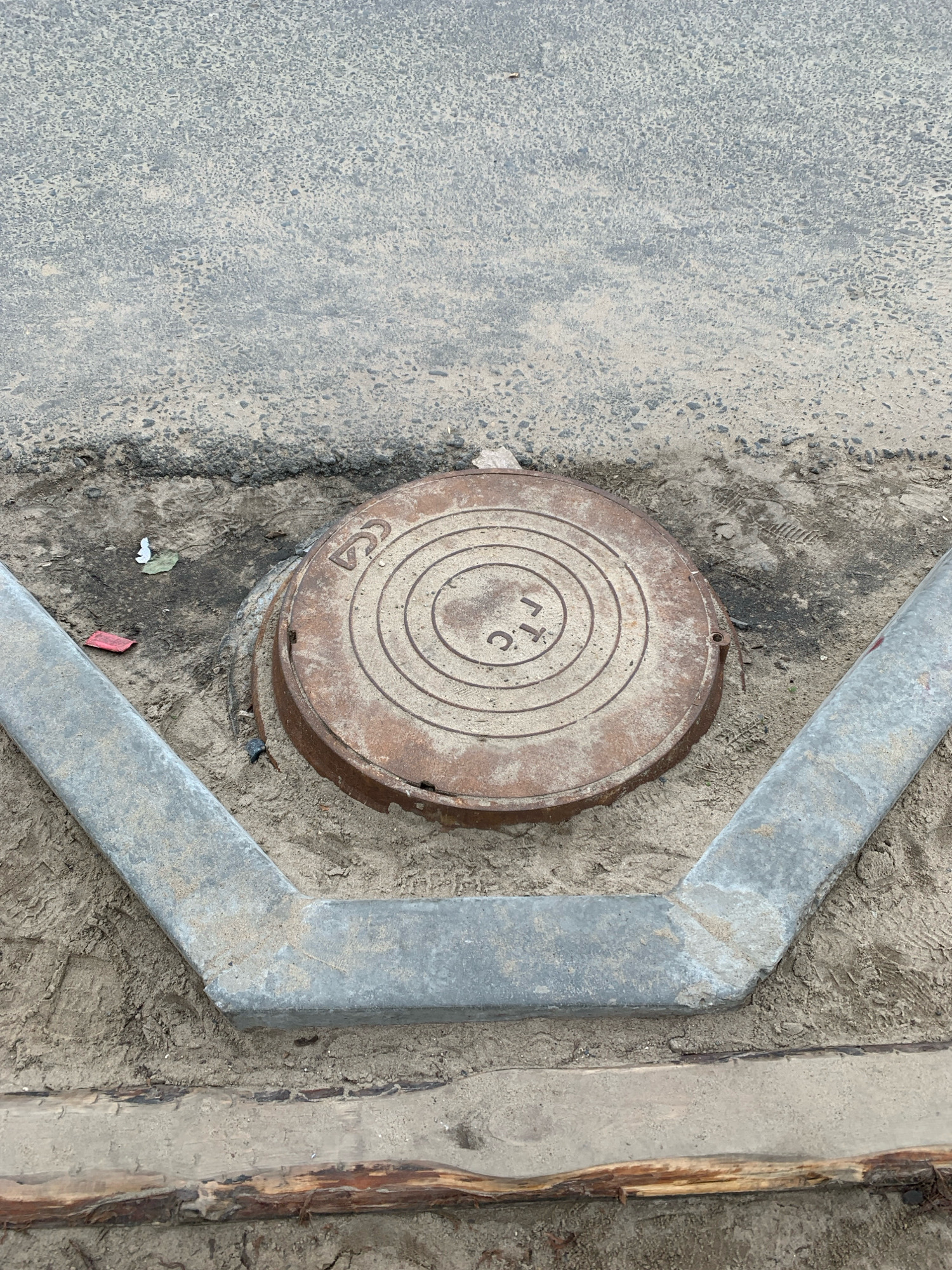 A rusted circular manhole cover with concentric rings and the Cyrillic letters "ГТС" is partially embedded in sandy soil. It is situated at the edge of an asphalt road, bordered by a V-shaped concrete curb. The scene appears to be an outdoor construction or repair site, with debris including a red wrapper and a piece of white paper scattered near the manhole. The environment is urban, likely within the city of Arkhangelsk, Russia, given the context.