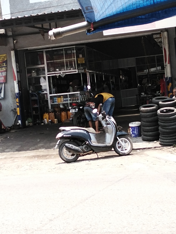 The image depicts an outdoor scene in Maron, Indonesia, in front of a motorcycle repair or tire shop under bright daylight. In the foreground, a dark grey and silver scooter, resembling a Honda Scoopy, is parked on its kickstand on a paved surface. Behind it, a male individual wearing a yellow shirt and blue jeans is bent over a dark motorcycle, appearing to work on it. Another individual, wearing blue shorts, stands near him, partially visible. Further to the right, an older male individual is seated, partially visible.
The shop entrance features a wide opening with a glass-paneled section on the left. Inside, shelves display various bottles and containers, likely automotive fluids. Several yellow jerrycans are visible on the floor inside the shop. A blue tarp awning extends over the entrance, with a grey drainpipe running beneath it. To the right of the entrance, multiple stacks of tires are arranged, alongside a blue and white bucket. The building itself has a grey facade with a corrugated metal roof extension. Partially visible signs, including "BM" and other unreadable text, are affixed to the storefront. The ground in the immediate foreground is a light-colored paved road or sidewalk.