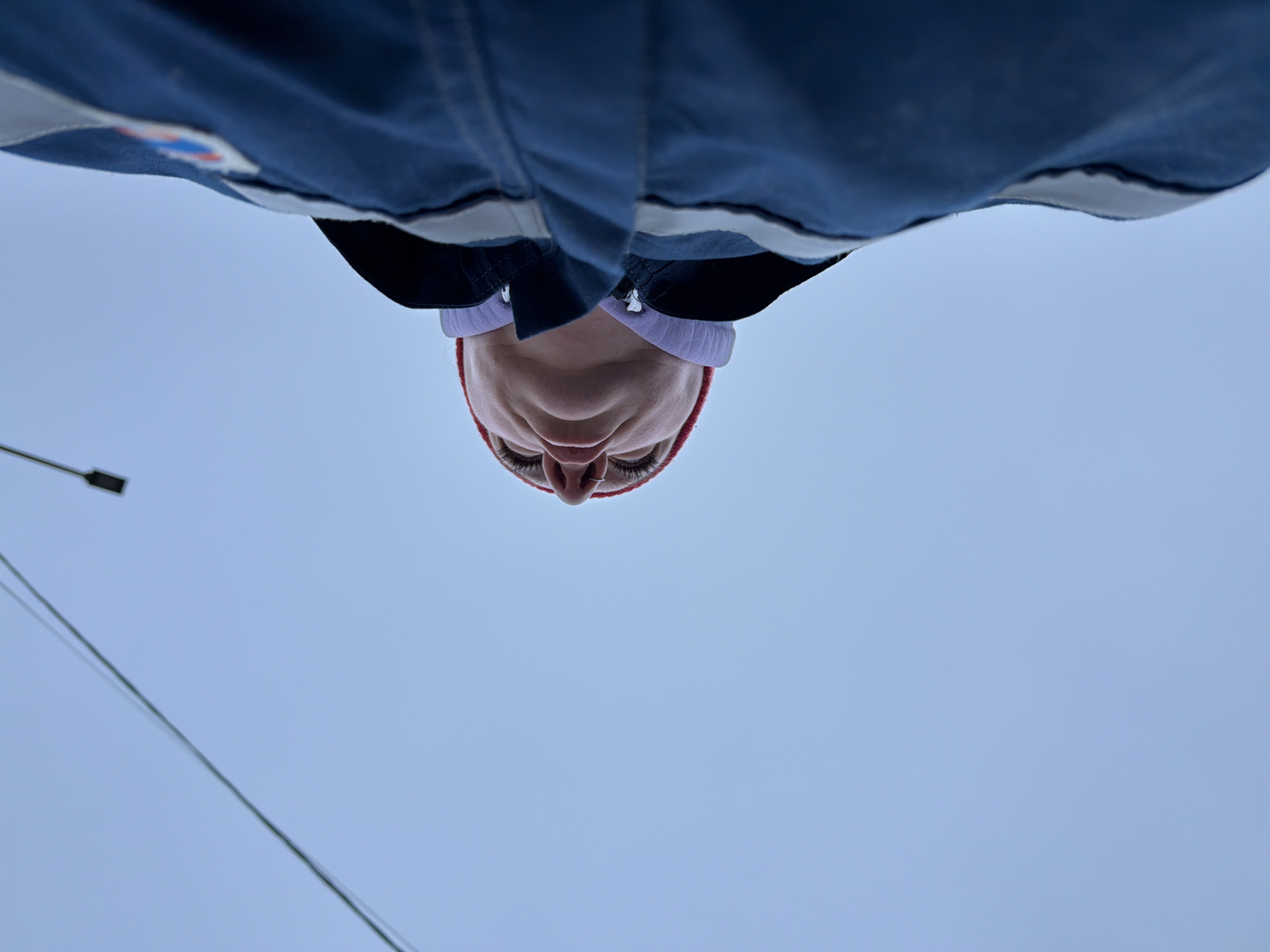An inverted, low-angle shot shows a person's face from below, framed against a pale blue sky. The person, possibly female, wears a red beanie and a purple headband. Part of a dark blue jacket with a white trim is visible around the upper part of the frame, suggesting the person is looking down at the camera. A thin wire or cable extends from the lower left corner towards the middle. In the upper left, a small black rectangular object is visible, likely a light fixture. The setting appears to be outdoors in an urban environment, potentially in Chaikovsky, Russia. No specific actions or interactions can be determined from the image.