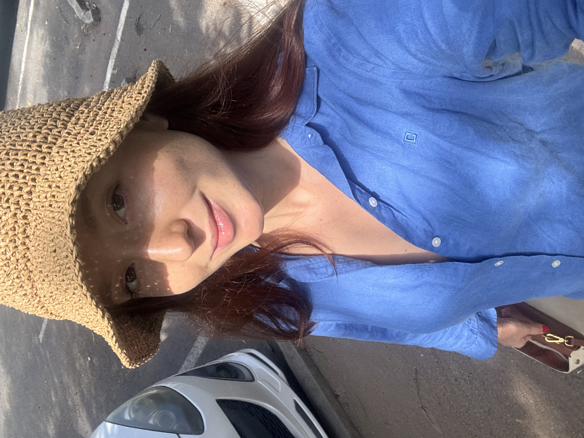 A fair-skinned woman with auburn hair, wearing a woven sunhat and a blue button-down shirt, poses for a selfie. She is positioned on a paved surface near a white vehicle. The location is indicated as Choeng Thale, Thailand.