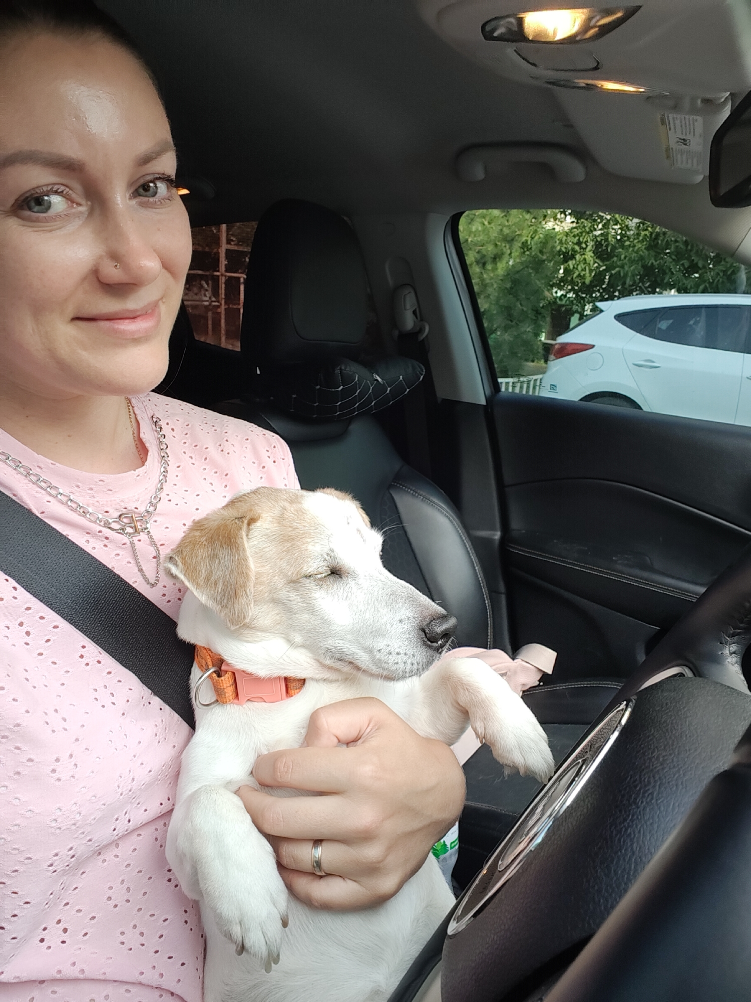 A woman is holding a small, white and tan dog in her lap while sitting in the driver's seat of a car. The dog, with its eyes closed, appears to be sleeping. The woman is wearing a pink, short-sleeved shirt with a cutout floral pattern and a silver necklace. She has a nose ring and her hair is pulled back. A seatbelt is visible across her chest. The interior of the car is black, with leather seats and a steering wheel. Outside the car window, trees and another white car are visible. The scene suggests it is daytime. The location is Chornomorsk, Ukraine.