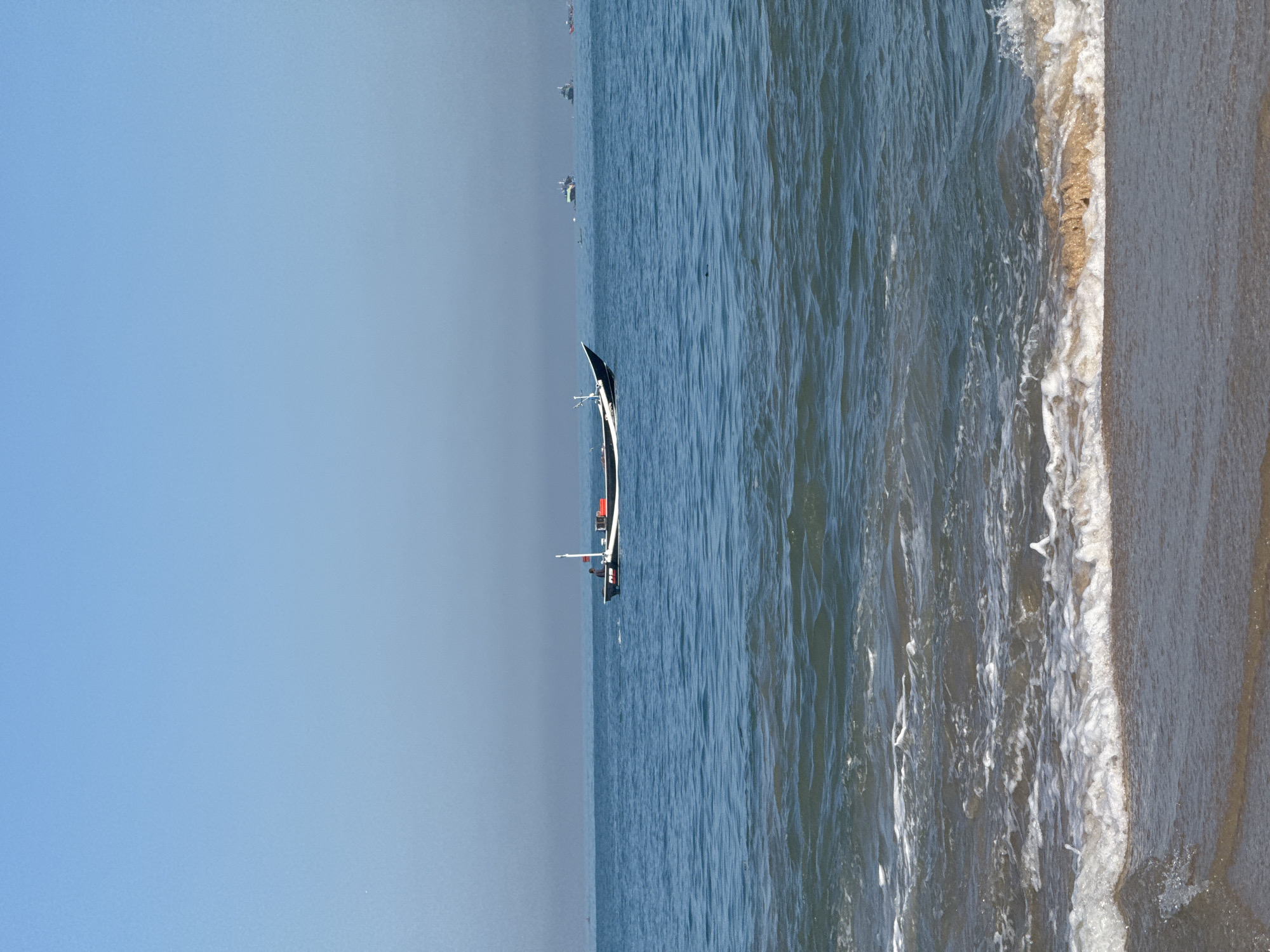 The image depicts a beach scene in Kuta, Indonesia, with a long, narrow boat floating in the water. The boat, white with a black sail-like structure, is positioned between the open sea and the shoreline. A person is visible in the boat. To the left of the boat, the sea stretches to the horizon under a clear, blue sky. To the right, waves gently wash onto the sandy beach. The scene suggests a calm day at sea, likely along the coast of Kuta.