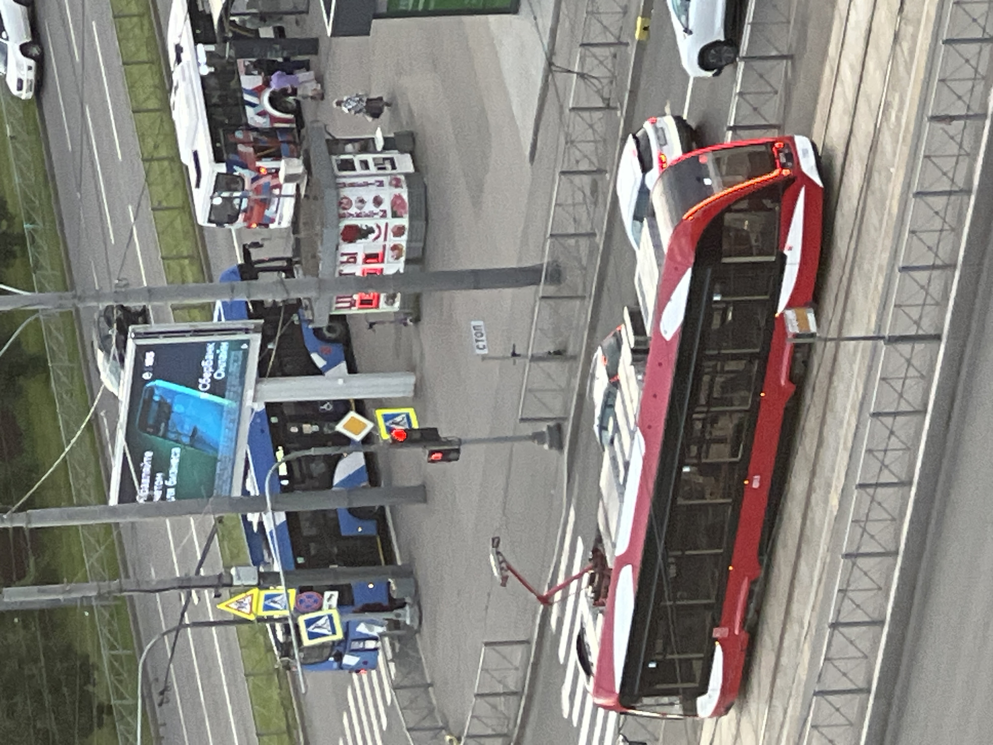 This image shows a view from above of a street scene in Sinyavino, Russia. The dominant subjects are several buses and cars. A bright red double-decker bus with white accents is prominently featured, driving on a tram track. Other vehicles, including a white car and a blue bus, are also visible on the road. The scene appears to be an urban or suburban environment with roads, sidewalks, and some greenery. There are traffic lights and various road signs, including diamond-shaped signs and signs with blue squares and white triangles.
The image seems to capture a moment of normal traffic flow. The lighting suggests it is daytime, possibly late afternoon or early evening, given the shadows. The weather appears to be fair.
Visible text includes "Сбербанк Онлайн" on a banner on the blue bus, which translates to "Sberbank Online," indicating a Russian bank. There is also a sign with the word "СТОП" (STOP) on a pole, and other illegible text on storefronts or advertisements.
The overall impression is that of a typical day in a Russian town with public transportation and vehicular traffic. There are no strong indications of a specific event, but rather a snapshot of daily life.