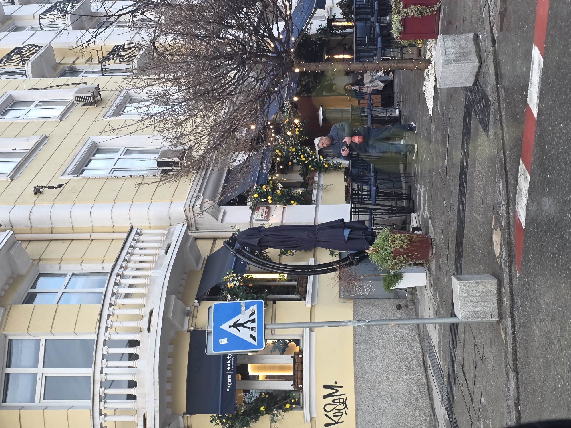 The image shows a street scene in Sofia, Bulgaria. A pedestrian crossing sign is visible. A man is standing outside a building, which appears to be a restaurant or cafe, decorated with flowers and string lights. He is holding a camera, possibly taking a photograph. Other people are seated at tables in the outdoor seating area. The building is light yellow with architectural details like a curved balcony. There is graffiti on the wall near the crosswalk.