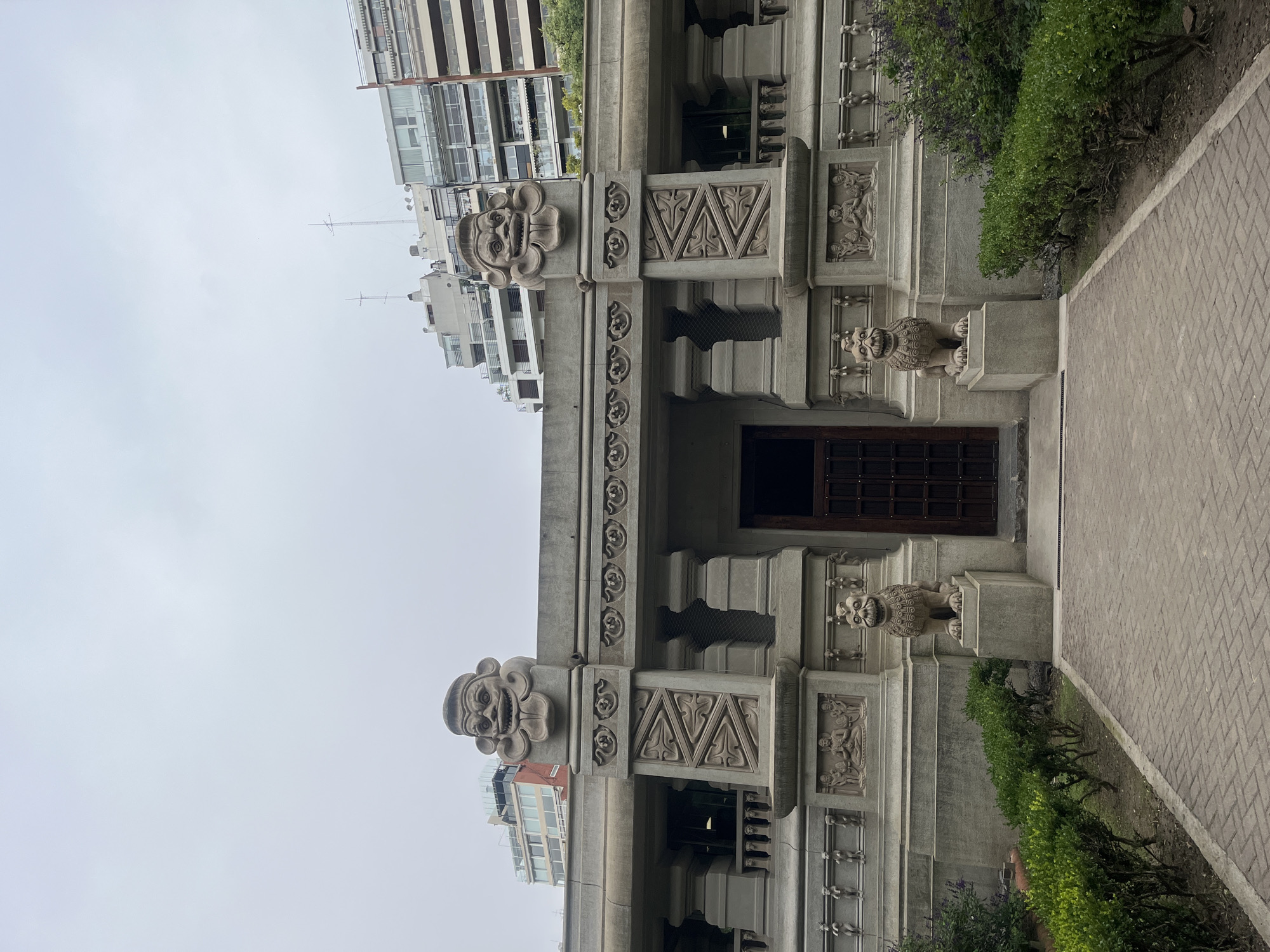 The image displays the facade of the Palacio Hindú (Ramakrishna Mission) located in Palermo, Buenos Aires, Argentina. The scene is static, devoid of any human presence or activity, under an overcast, light grey sky.
The central subject is a multi-layered, grey stone building exhibiting Indo-Saracenic architectural characteristics. A dark, double-leaf wooden door serves as the central entrance at ground level. This entrance is flanked by two identically sculpted, seated stone lion statues, each positioned on a rectangular stone plinth. A herringbone-patterned brick pathway leads directly to the entrance, bordered on both sides by manicured green hedges and clusters of purple-flowering shrubs planted in bare earth.
Above the entrance, the facade is extensively decorated with stone carvings. Two prominent, stylized, mask-like faces with wide grins and protruding tongues adorn the upper corners of the main building structure, surmounting decorative pillars. Below these, the facade incorporates a series of bas-reliefs depicting geometric patterns, intricate floral motifs, and figures, including some seated in meditative postures and others standing. Windows with dark grates are visible behind balustrades composed of small stone columns.
In the background, rising above the Palacio Hindú, several modern, multi-story residential apartment buildings with numerous balconies and rooftop antennas are visible, indicating an urban environment.