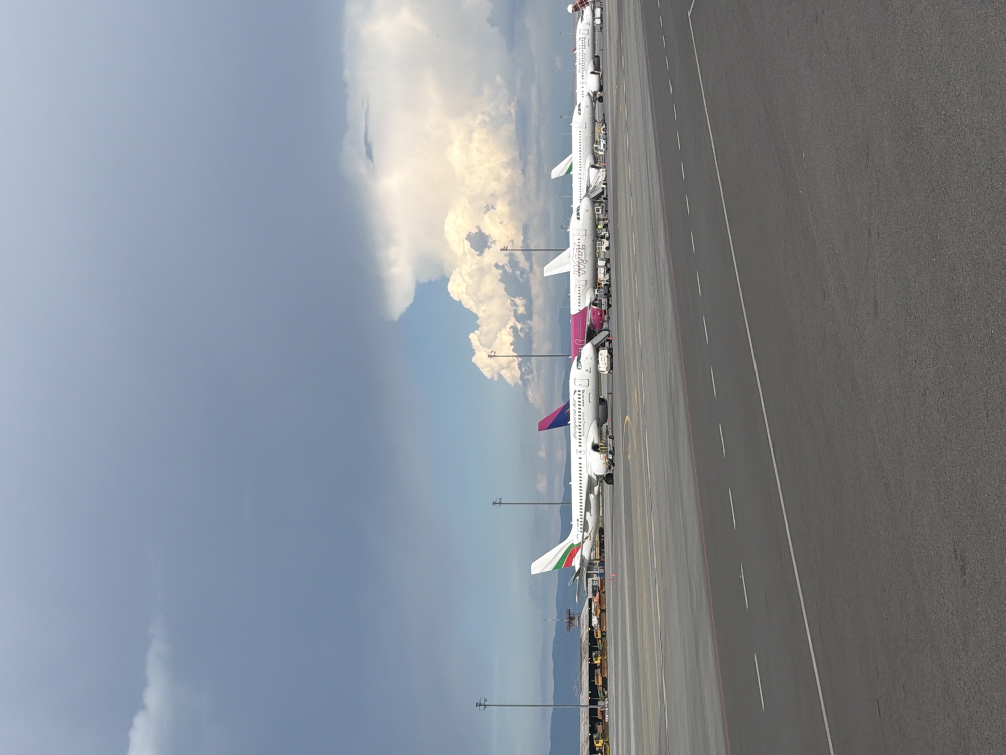 This media file captures an exterior view of an airport tarmac on a cloudy day, with a line of parked airplanes. The scene is set at Sofia Airport in Bulgaria. Several commercial airplanes are visible, lined up on the tarmac, likely awaiting or having just completed passenger boarding or deplaning. The sky is a mixture of gray clouds and brighter, sunlit clouds, suggesting dynamic weather conditions, possibly transitioning between overcast and sunny periods. The ground is asphalt with clear lane markings.
Notable details include the distinct tail fins of some of the aircraft, one featuring the colors of the Bulgarian flag (white, green, and red), and another with a purple and blue design. Some text is visible on the side of one aircraft, appearing to be airline branding. The lighting and cloud formations suggest it is daytime, possibly late morning or afternoon. The overall impression is one of a busy, functional airport environment.