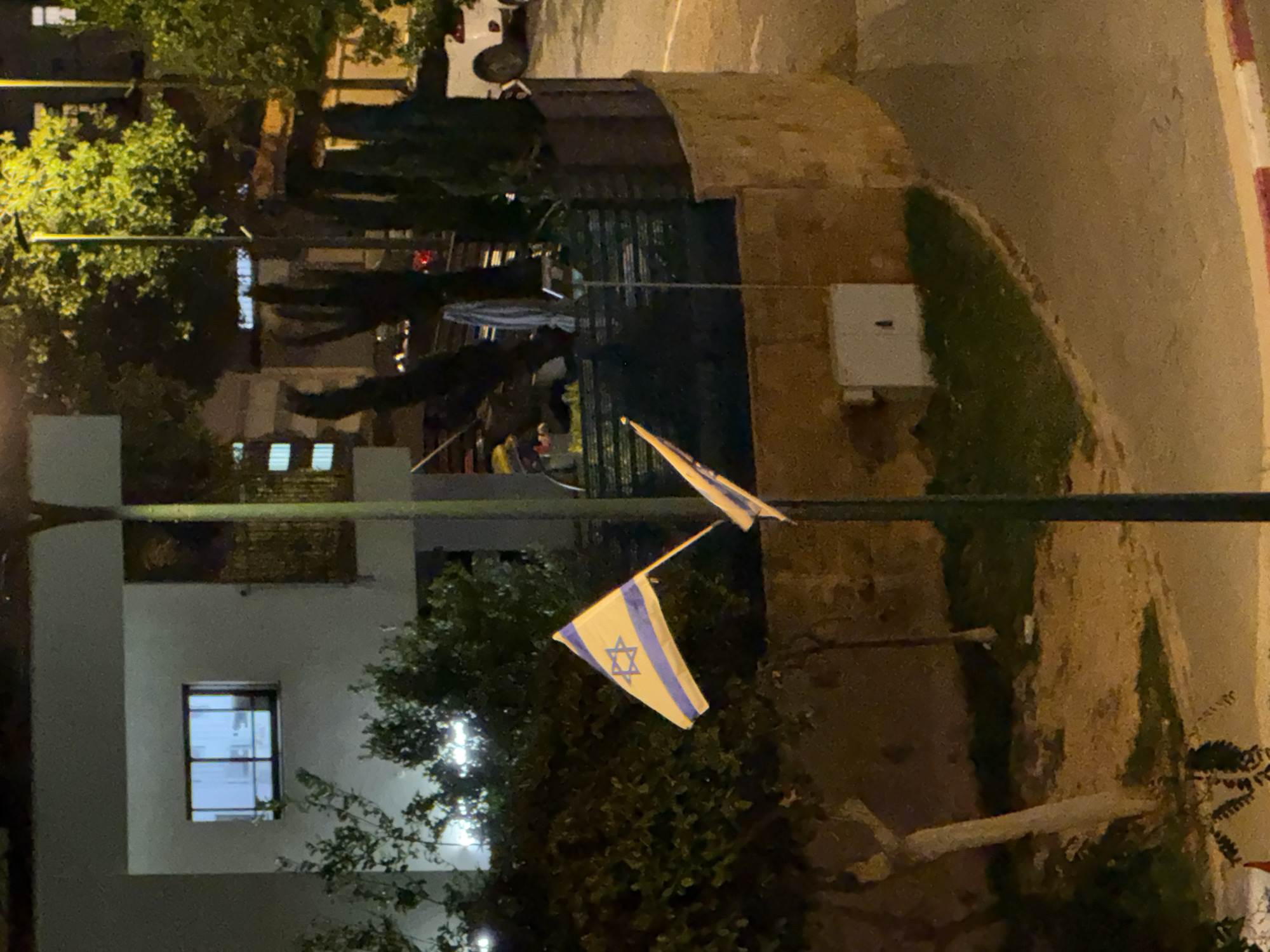 A night view in an unknown city shows two Israeli flags. They are attached to a vertical pole with a horizontal arm at the top. In the background, there is a light-colored building with a window and trees. A street and a parking lot are also visible.