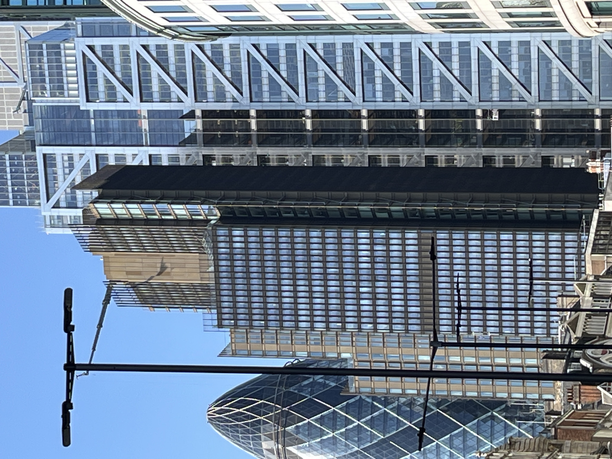 This is a photograph of the City of London skyline. The image is dominated by modern skyscrapers, including the iconic Gherkin building on the left. The Gherkin is a distinctive glass skyscraper with a rounded, conical shape. To its right, a dark, rectangular skyscraper stands tall, its facade made up of numerous windows. Further to the right, a large building with a striking geometric, metallic exoskeleton is visible. This building features a criss-cross pattern of diagonal beams against a backdrop of glass windows. The sky above is a clear, bright blue. In the foreground, silhouetted against the buildings, are several black lampposts with multiple lights. The lower part of the image shows glimpses of older, traditional architecture with brickwork and arched windows, suggesting a mix of historical and contemporary urban development. There are no people visible in the frame.