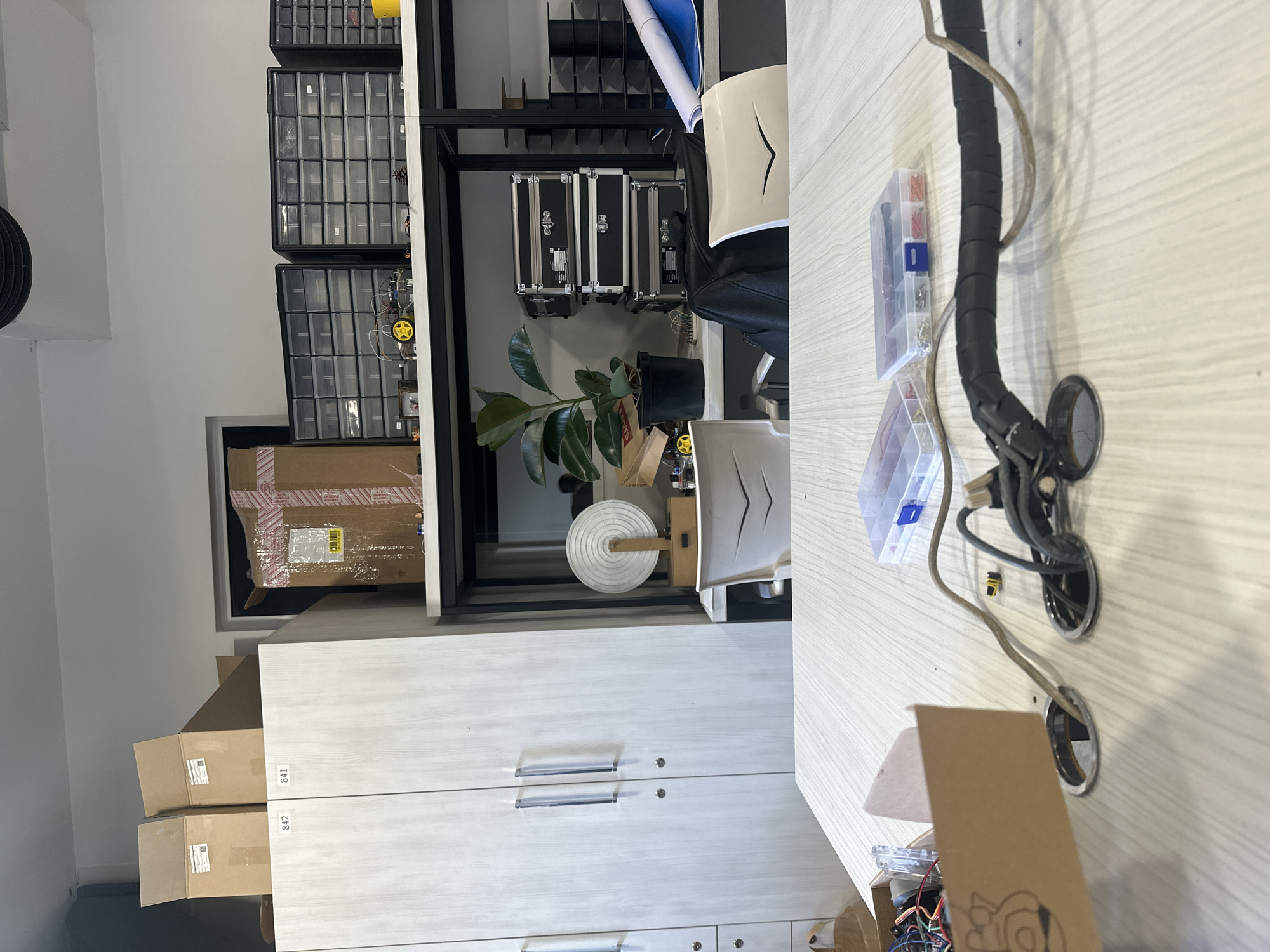 A cluttered indoor workspace is depicted. The scene appears to be a workshop or hobby area.
**Objects:**
* A cabinet with multiple drawers.
* Several cardboard boxes stacked near the cabinet. Some have labels.
* A metal shelving unit.
* Storage drawers with compartments in the shelving unit.
* Three black cases with metal edges in the shelving unit.
* A green plant in a black pot.
* A white disc with a wooden rod on the shelving unit.
* Two desk chairs with white and black elements.
* A desk with various items including a black cable conduit, electrical connectors in clear plastic cases, and an unfinished project in a cardboard box.
**Environment:**
* The walls are painted white.
* The lighting appears to be artificial.
* There is a sense of disorder and a lot of projects in progress.
**Location:**
The location cannot be identified as Yerevan, Armenia based on the image provided. There are no visual cues or indicators that explicitly place the scene in that city.