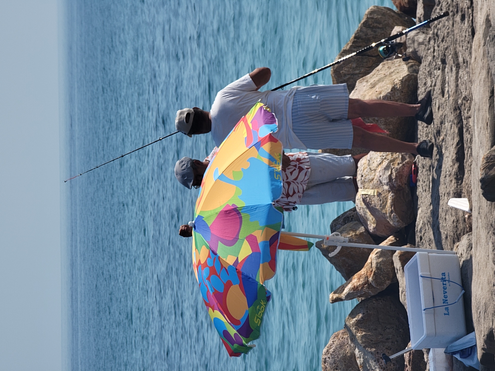Two people are fishing on a rocky shoreline. The person in the foreground is partially obscured by a colorful umbrella with a floral pattern. They are wearing a white shirt and striped shorts. The person behind them is wearing a white t-shirt and light-colored shorts. Both appear to be casting or retrieving fishing lines, as fishing rods are visible. The scene is set against a backdrop of calm blue water and large, rough rocks. The lighting suggests it is daytime, likely sunny. A white cooler with "La Neverita" written on it is visible on the rocks. The activity appears to be recreational fishing.