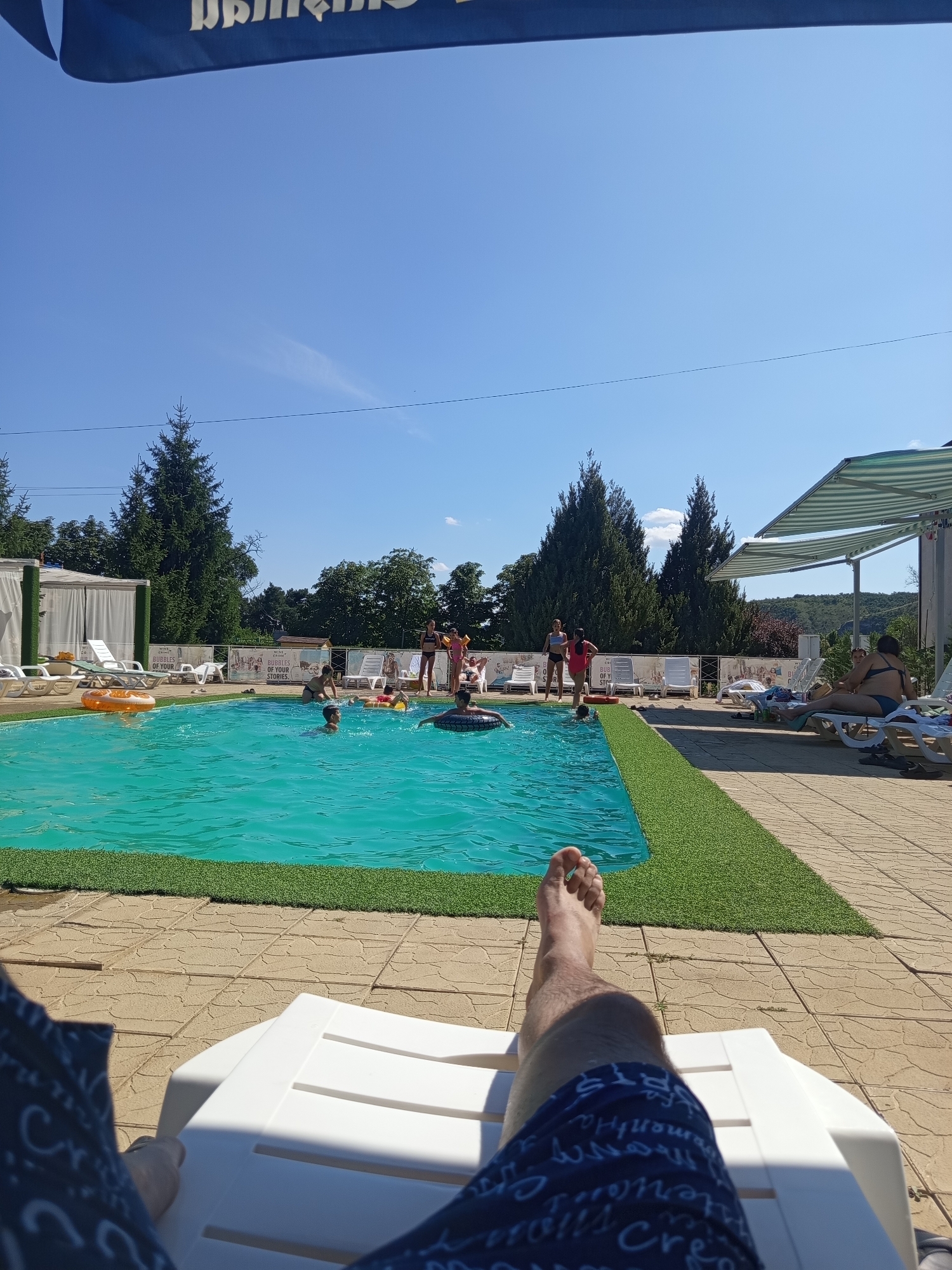 The image depicts a sunny day at an outdoor swimming pool facility, possibly in Molovata Nouă, Moldova. In the foreground, a person's legs and feet are visible, resting on a white lounge chair. The person is wearing blue patterned shorts. The lounge chair is positioned on a paved patio area with a textured tile pattern. Adjacent to the patio is a bright turquoise swimming pool, with several people swimming and playing. Some individuals are using inflatable rings. Green artificial turf lines the edge of the pool. In the background, white tents or cabanas are set up, and further beyond, lush green trees and a clear blue sky are visible. A striped awning provides shade over some of the seating area. The overall scene conveys a leisurely summer atmosphere.