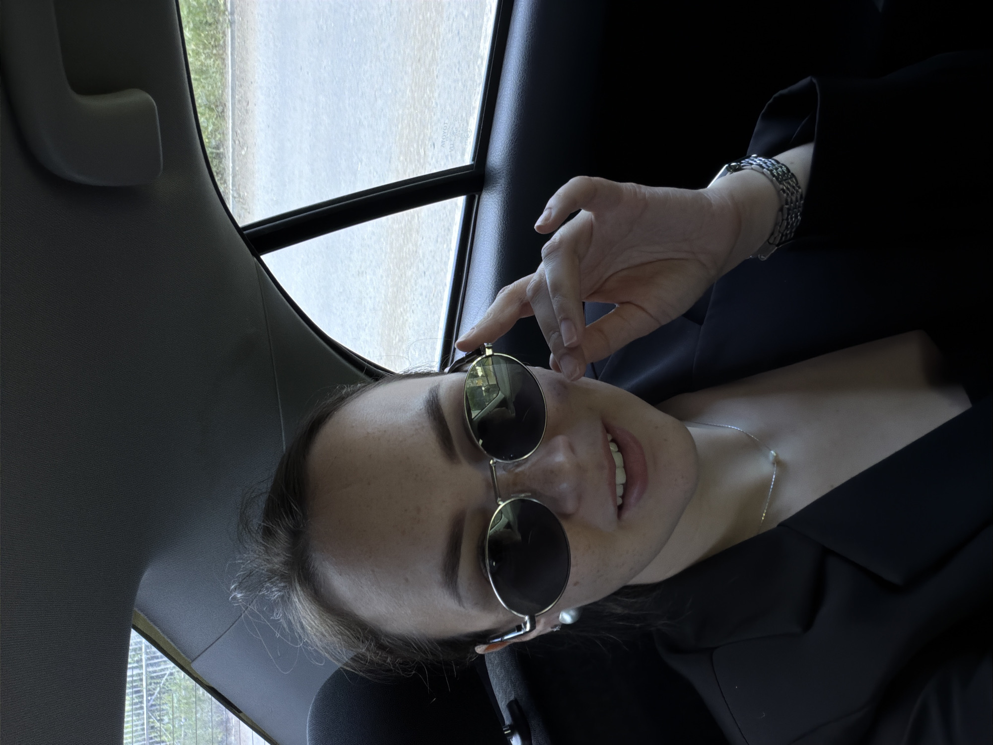 A young woman with light brown hair pulled back is seated in the back of a car, looking towards the camera. She is wearing a black blazer, a silver necklace with a pearl pendant, and round, silver-rimmed sunglasses. She has a small smile on her face and her right hand is near her face, adjusting her sunglasses. The interior of the car, including the headliner and a portion of the rear window, are visible. Through the rear window, trees and a road can be seen, indicating an outdoor environment, possibly a suburban or rural area on the outskirts of Chaikovsky, Russia.