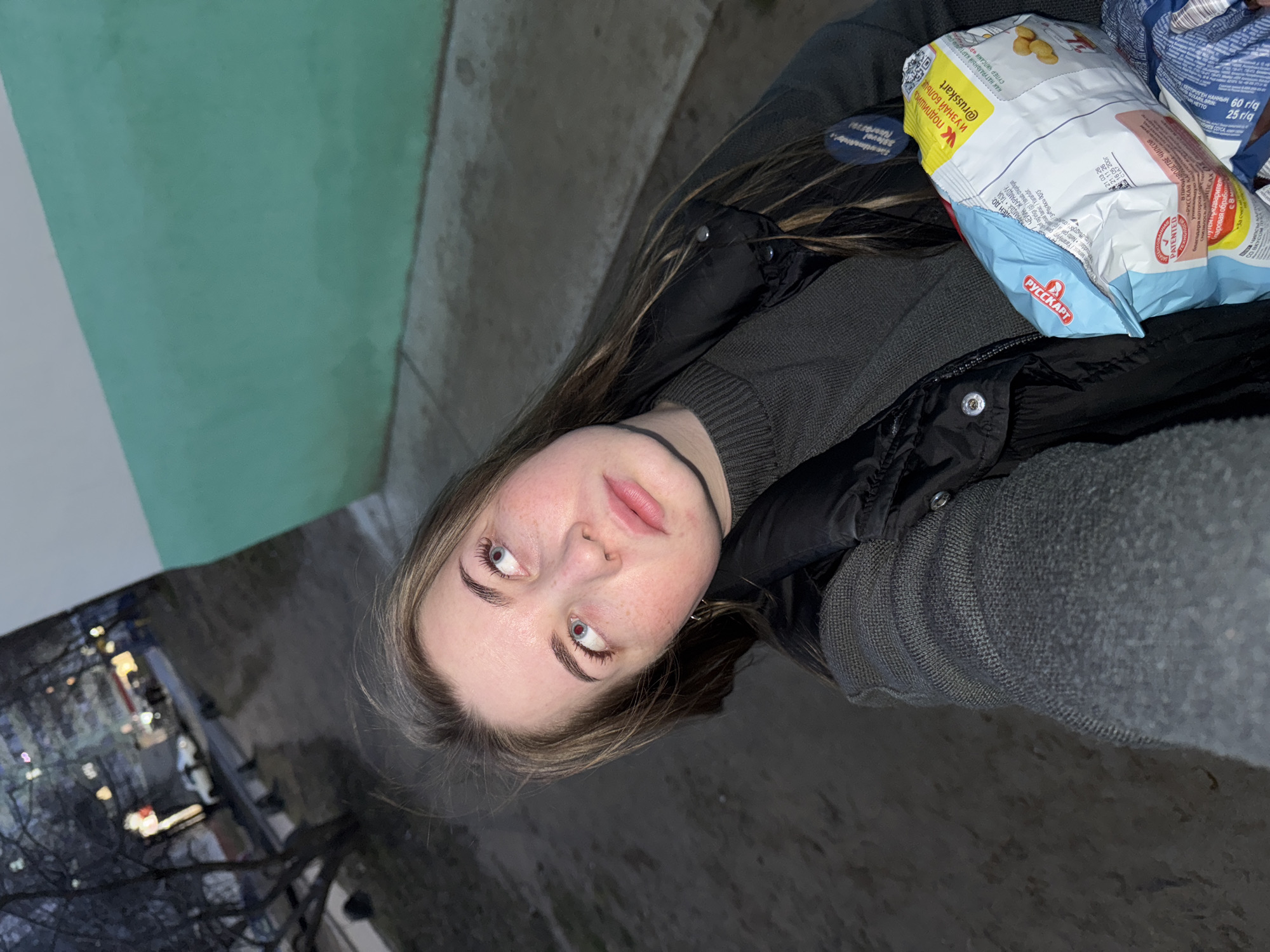 The image is a close-up, slightly angled shot of a young woman. She has fair skin with freckles, light eyebrows and eyelashes, and her eyes are directed slightly upward and to the left. She wears a dark choker around her neck.
She is wearing a gray sweater under a black puffer vest or jacket. A bag of chips with Russian text, including "@russkart," rests on her chest.
The background is blurry and includes a muted green and white wall on the left. On the left, there's a nighttime urban scene with lights and bare tree branches. The image appears to be taken at dusk or night. The chips contain some text in the Russian language. Based on the OCR of the image, the chips are called "РУССКАРТ," which also is a brand. I cannot verify the location.