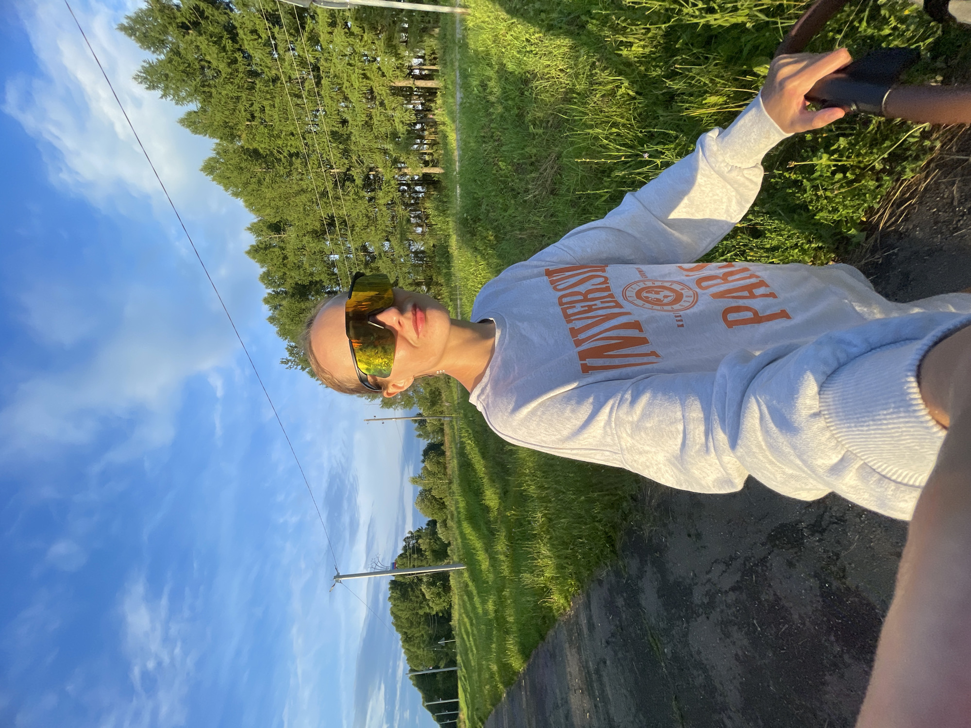 A woman wearing oversized reflective sunglasses and a gray sweatshirt with orange lettering is standing outside on a path. The sweatshirt has the word "UNIVERSITY" across the chest, and below that, "PARIS." She is holding onto the handle of a stroller or bicycle. The background features a field of tall green grass, a line of evergreen trees, and telephone poles with wires running through them. The sky is blue with scattered white clouds, and the sun appears to be setting, casting long shadows. The location is the city of Podshivalovo, Russia.