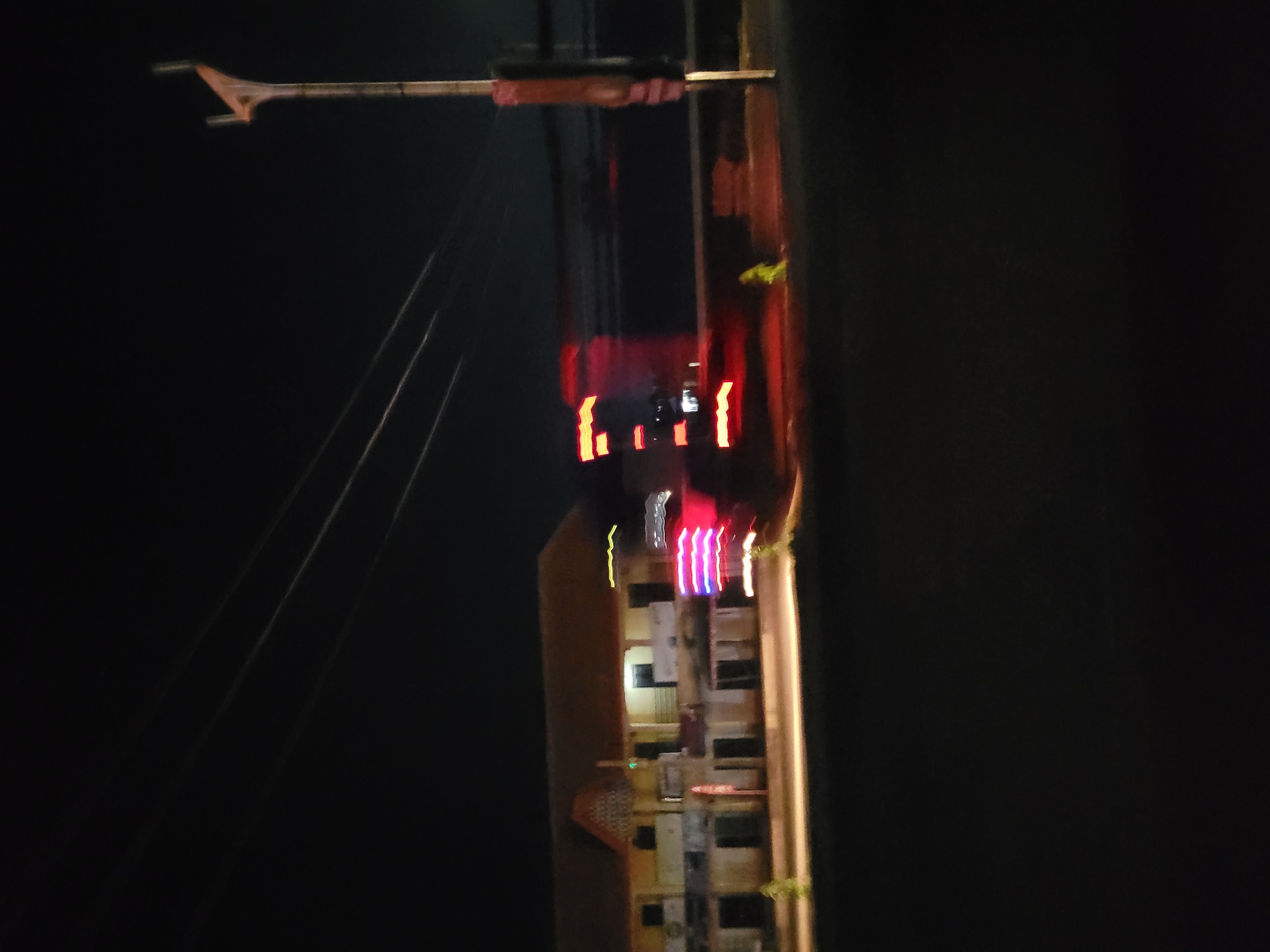 The image shows a vertical view of a dark city street at night. There are street lights and traffic lights illuminating sections of the buildings, with many of the lights out. There is an antenna on a tall pole and overhead power cables extending from it. The location is Unknown, Unknown.