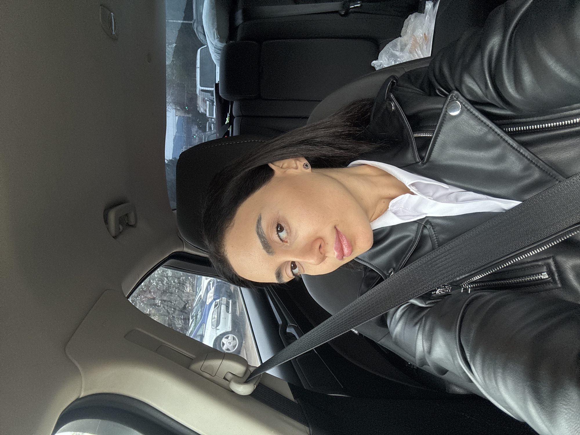 A woman is seated in a vehicle, wearing a black leather jacket over a white shirt and a fastened seatbelt. The photo is taken from inside the car, looking towards the woman. Outside the car window, vehicles are visible suggesting a city street. Based on the street conditions and architectural style, the location is likely Tbilisi, Georgia.