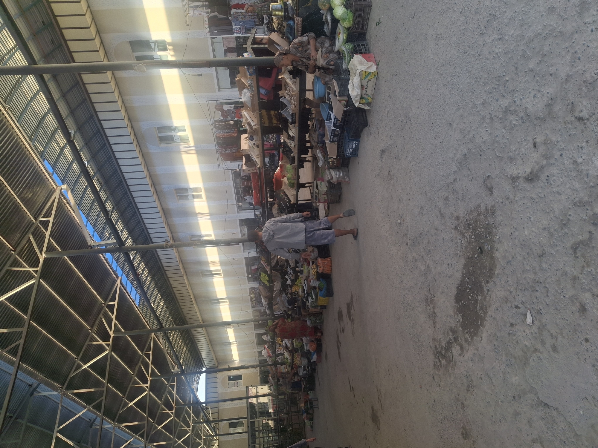 The image depicts an outdoor marketplace, likely in Ko`kdala, Uzbekistan, under a covered structure with a corrugated metal roof supported by metal beams and columns. Sunlight streams through gaps in the roof, casting long shadows across the scene. The ground is made of compacted dirt and concrete, with some wet patches and debris.
In the foreground, a man wearing a grey hoodie and blue shorts walks away from the camera. To the right, a woman in a patterned dress and headscarf is seated behind a table laden with produce. Various fruits and vegetables, including watermelons and cabbages, are displayed in crates and on the ground. Stalls with wooden shelves are visible, stocked with more goods. In the background, more market stalls and buildings with windows are present. Clothing and other items hang from racks. Other individuals can be seen browsing or working at the stalls, though they are less distinct. The overall impression is a busy, open-air market with a variety of goods for sale.
