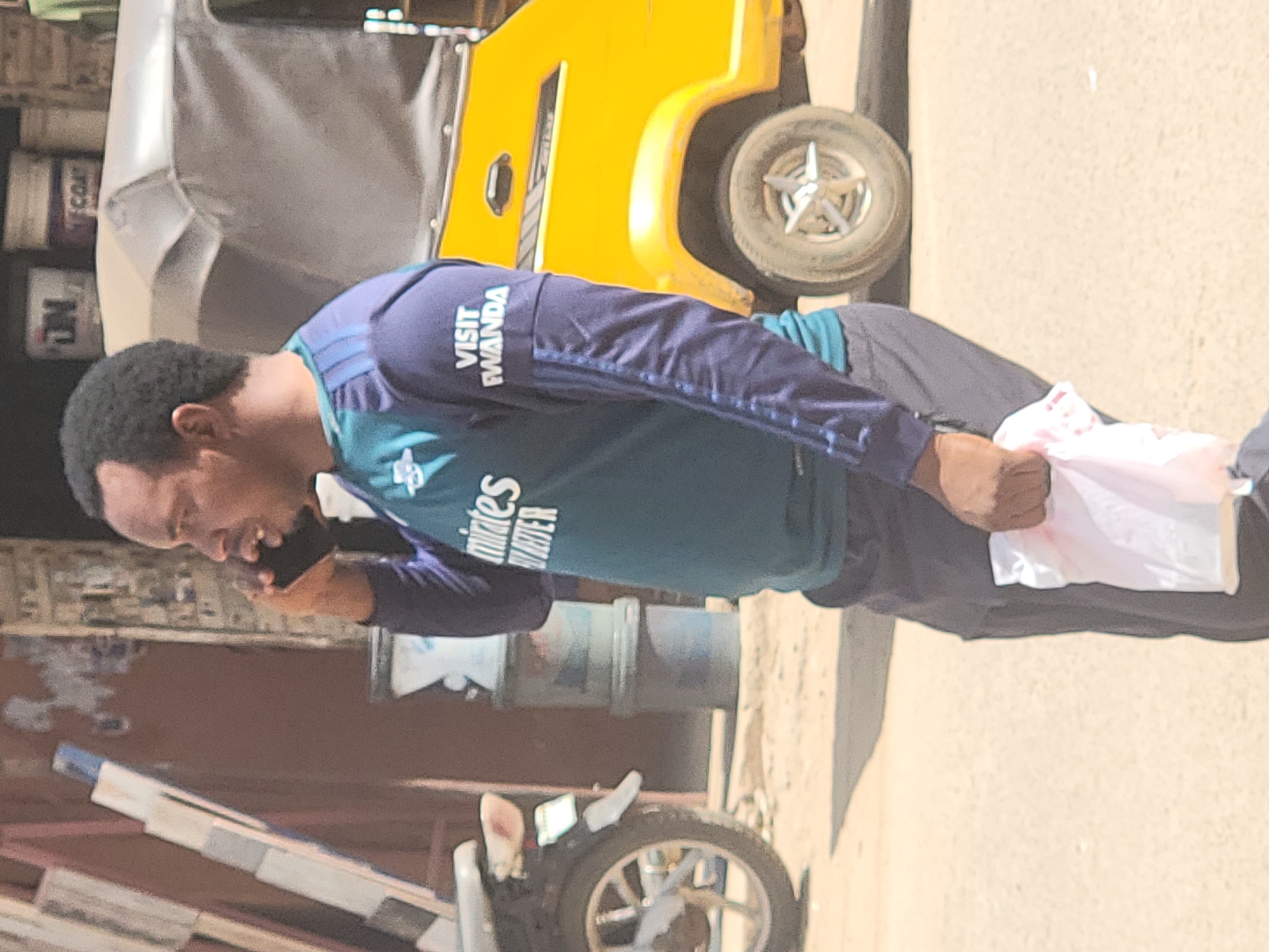 A man with short black hair is standing on a dusty street, holding a white plastic bag and talking on a mobile phone. He is wearing a long-sleeved shirt with the words "VISIT RWANDA" on the sleeve and "Emirates" on the front. Behind him, there is a yellow three-wheeled vehicle and various objects, including some containers and building materials. The location appears to be a bustling urban area, likely in Maiburiji, Nigeria.