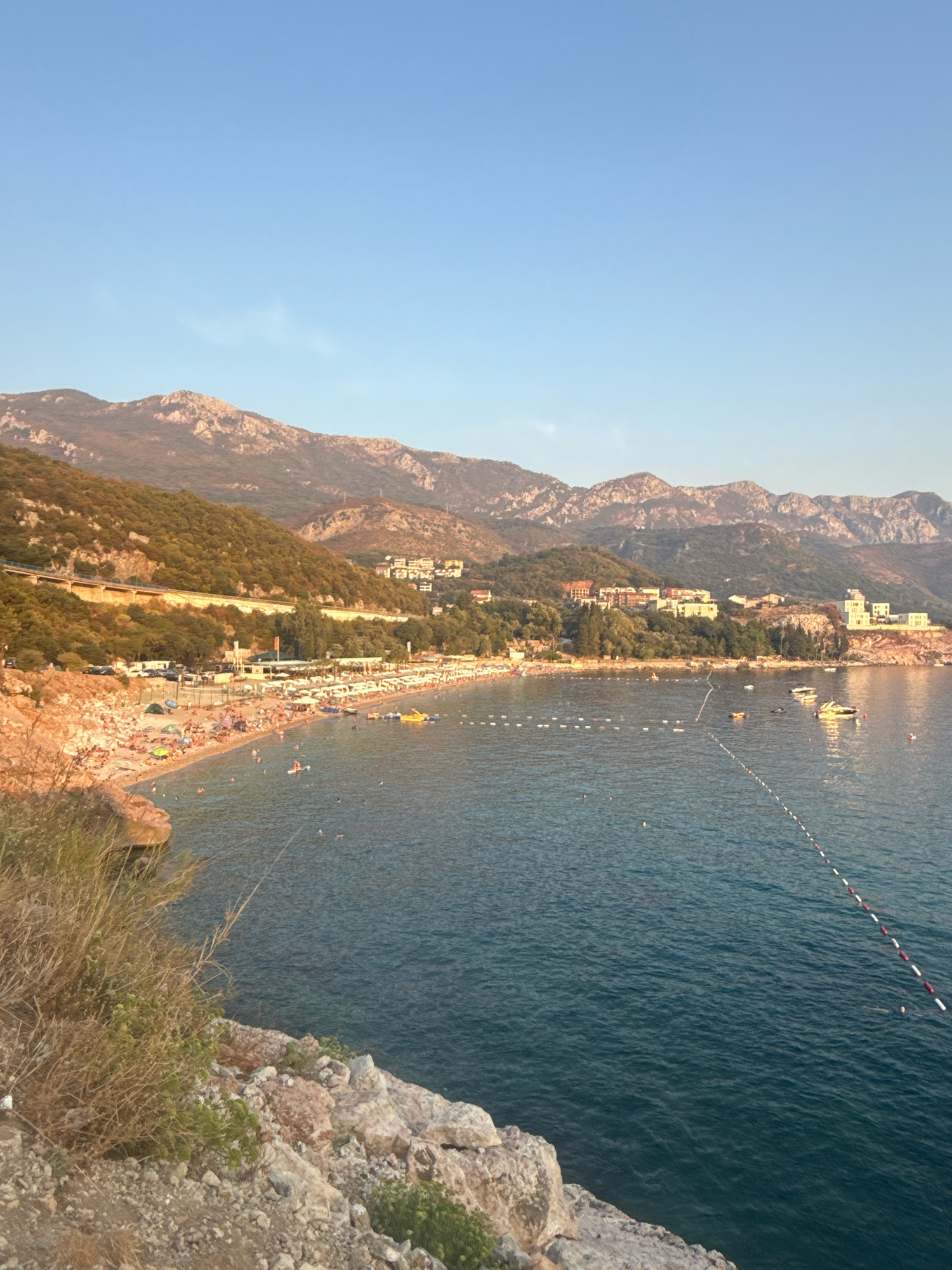 The image shows a wide view of a beach and the sea in Kamenovo, Montenegro. The sky is clear and blue. In the background, there are rolling hills and mountains covered in trees. On the hillside above the beach, there are several buildings, some appearing to be hotels or residences.
The beach itself is quite crowded with many people sunbathing, walking, and swimming. There are numerous white umbrellas and lounge chairs set up on the sand. A road runs along the top of the beach area, with cars parked along its side. Boats are visible in the water, including several white motorboats and some smaller vessels. A rope with buoys demarcates an area of the water. The sea is a deep blue color, with gentle waves. In the foreground, there are rocky areas and some dry vegetation.