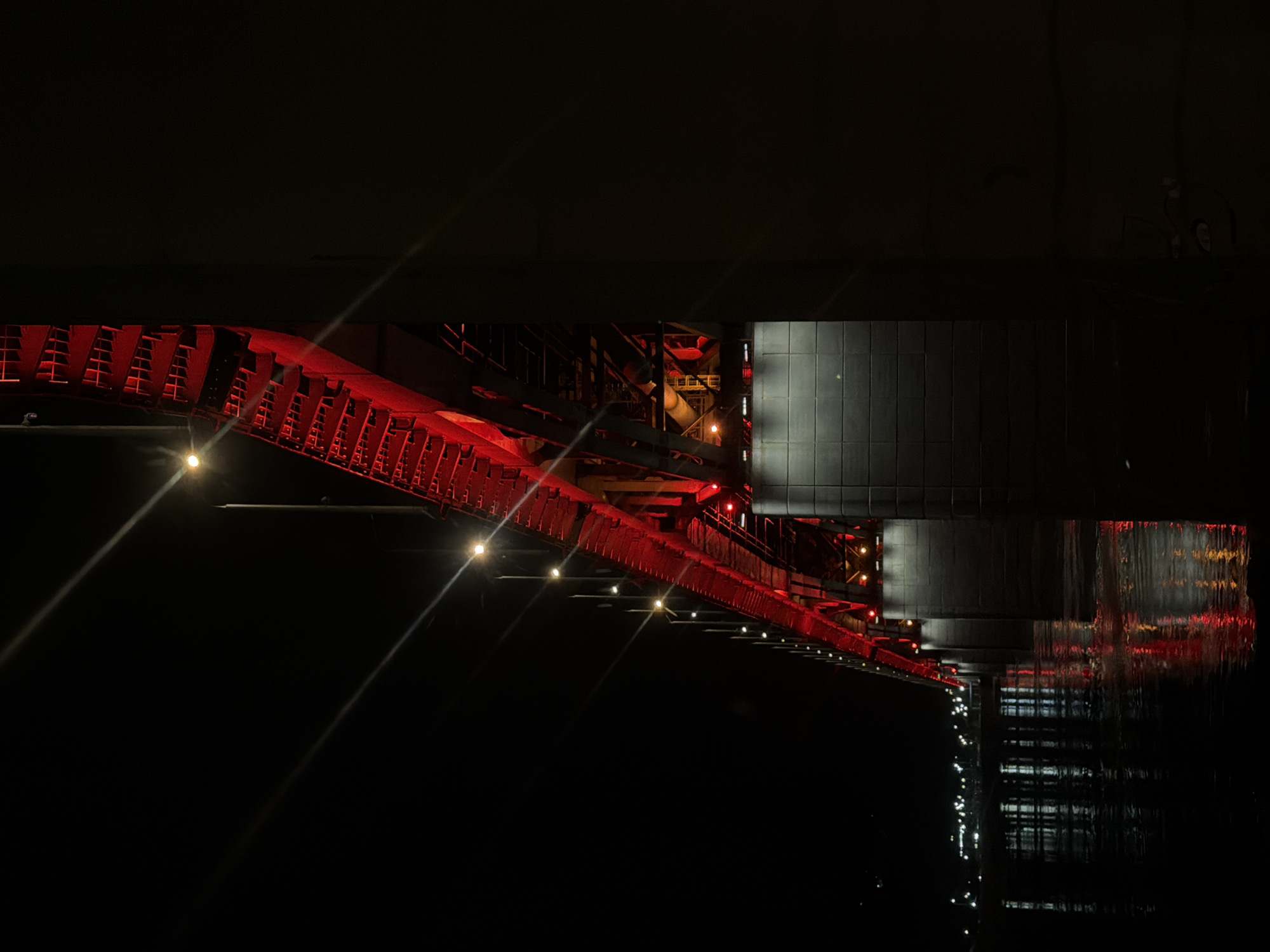 The image captures a dimly lit, nighttime view of the Vinogradovsky Bridge in Krasnoyarsk, Russia, as seen from below. The bridge's red structural elements, including latticework and supports, are partially visible against the dark background. Sparse, bright lights line the underside of the bridge, creating distinct light streaks. A section of the bridge's supporting structure, appearing as a dark, tiled wall, is present towards the right. Reflected light and colors are distorted on the water's surface below. The overall scene is characterized by darkness and selective illumination, highlighting the bridge's architecture.