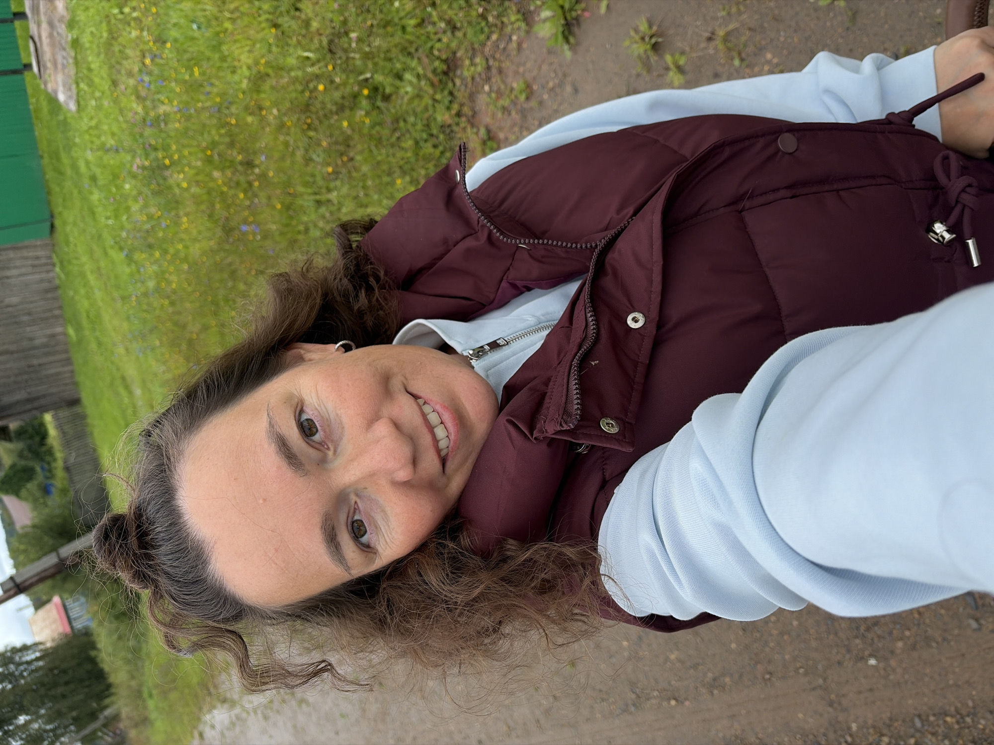 A woman with curly brown hair tied in a bun and a silver hoop earring smiles at the camera. She is wearing a light blue hoodie and a burgundy puffer vest. The background appears to be a rural area with a dirt road, green grass, and some scattered flowers. In the distance, there are wooden structures and a green fence. The image seems to be a selfie taken outdoors. The location is in the village of Uzier, Russia.