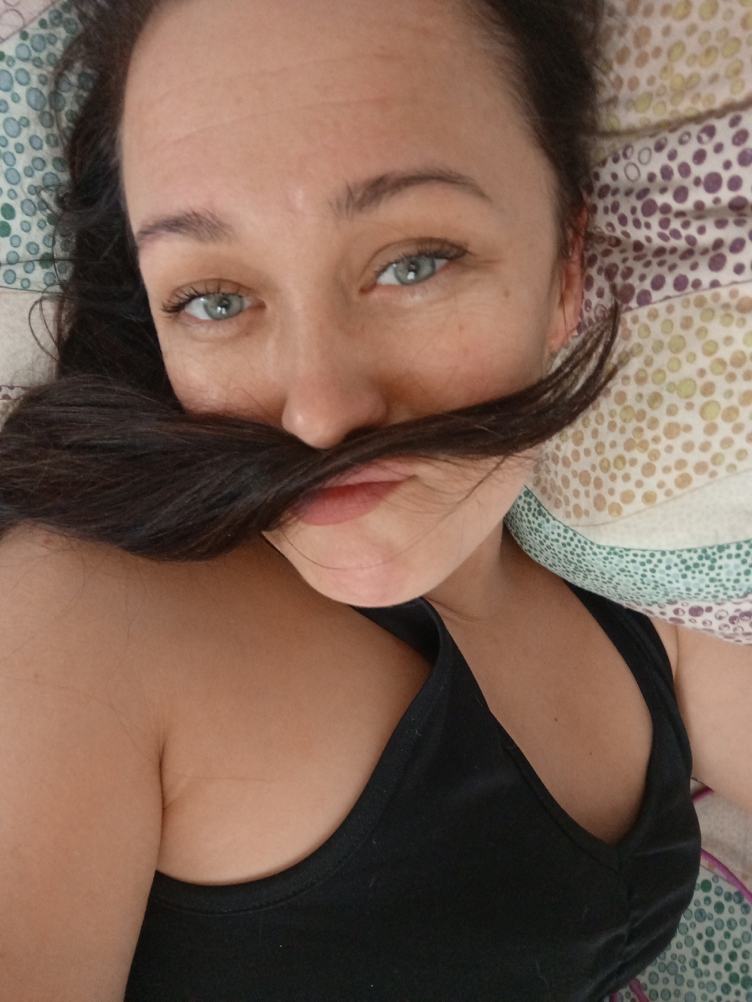 The image captures a close-up of a woman lying down, looking directly into the camera with a playful expression. She has light blue or green eyes and dark brown hair, a section of which she holds across her upper lip to mimic a mustache. Her lips are slightly parted in a subtle smile. She wears a plain black tank top or camisole, visible on her upper body.
The setting appears to be indoors, likely a bedroom, indicated by the patterned bedding around her head and shoulders. On the left, the bedding features a light background with green and blue circular patterns. On the right, the bedding has a light background adorned with purple, yellow, and beige irregular polka dot patterns. The lighting suggests a bright indoor environment, possibly daytime. The woman's posture is relaxed, and her gaze is direct, conveying a candid and lighthearted moment. There is no visible text in the image. The provided location context is Chaikovsky, Russia.