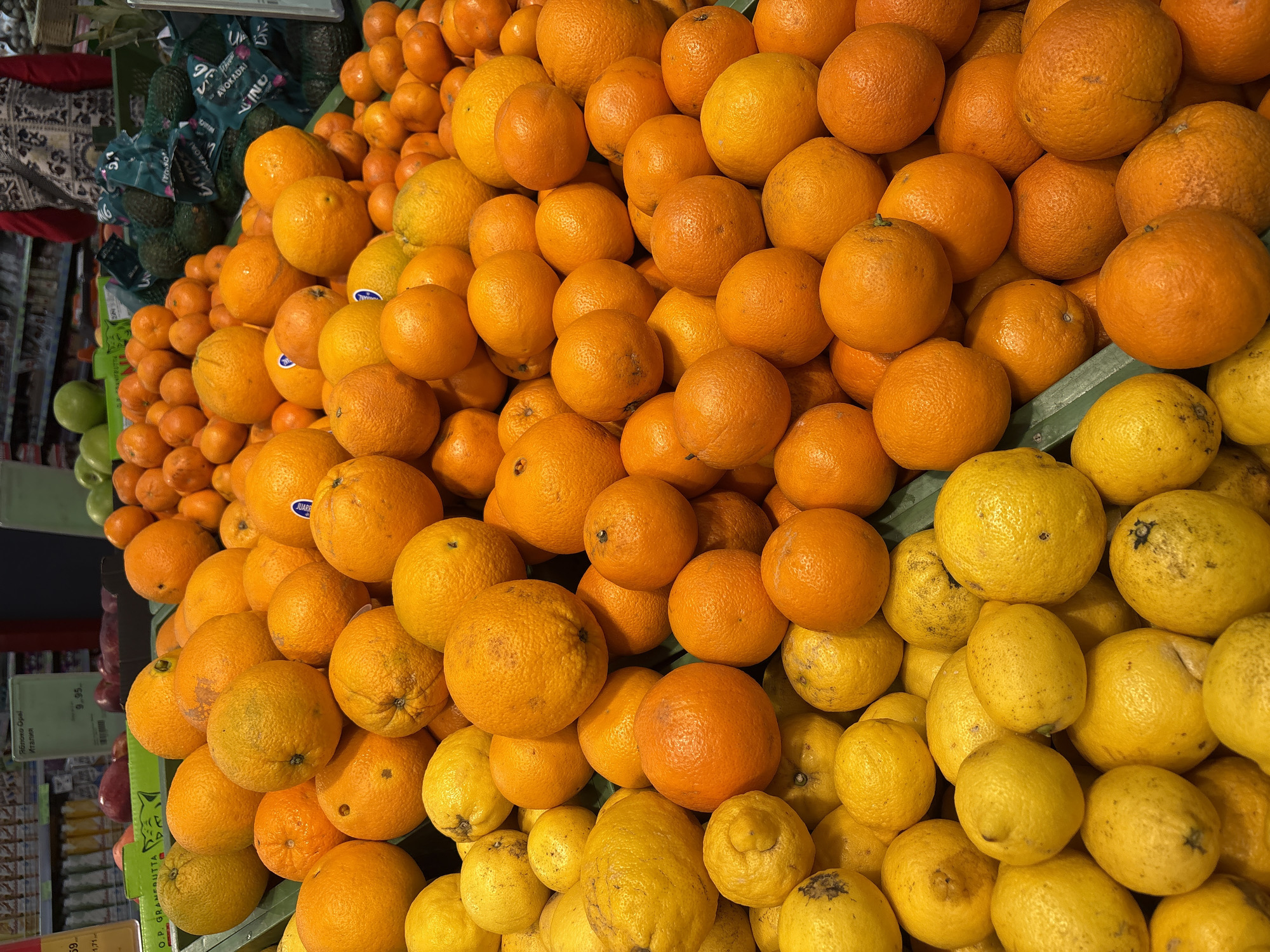 A grocery store display in Minsk, Belarus, shows a pile of oranges and lemons. The oranges are various sizes, with some smaller mandarin oranges mixed in. At the bottom right, a section of yellow lemons is visible. To the left of the oranges, there are green apples, avocados in branded bags and signage related to pricing. The display is brightly lit.