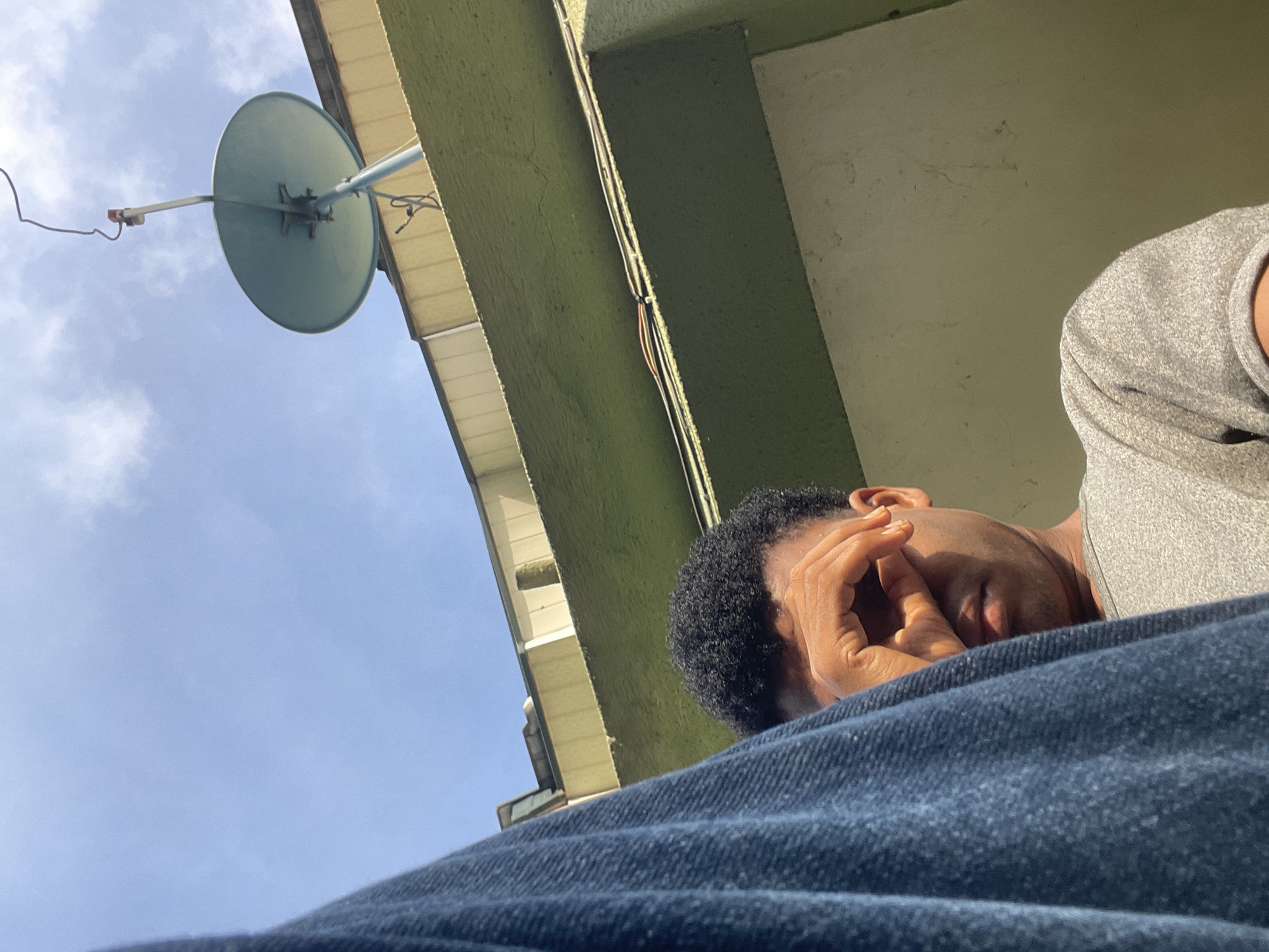 The image is taken from a low angle, looking upwards at a man lying down outdoors. The man has dark hair and brown skin, wearing a gray shirt and dark denim pants. His right hand is raised to his face, partially obscuring his features.
Overhead, the eaves of a building are visible, painted green and cream. A satellite dish is mounted on the eaves, its light blue-grey surface contrasting with the sky. The sky is a pale blue with light clouds.
The location is in Ado, Nigeria. This is based on the geographical location given.