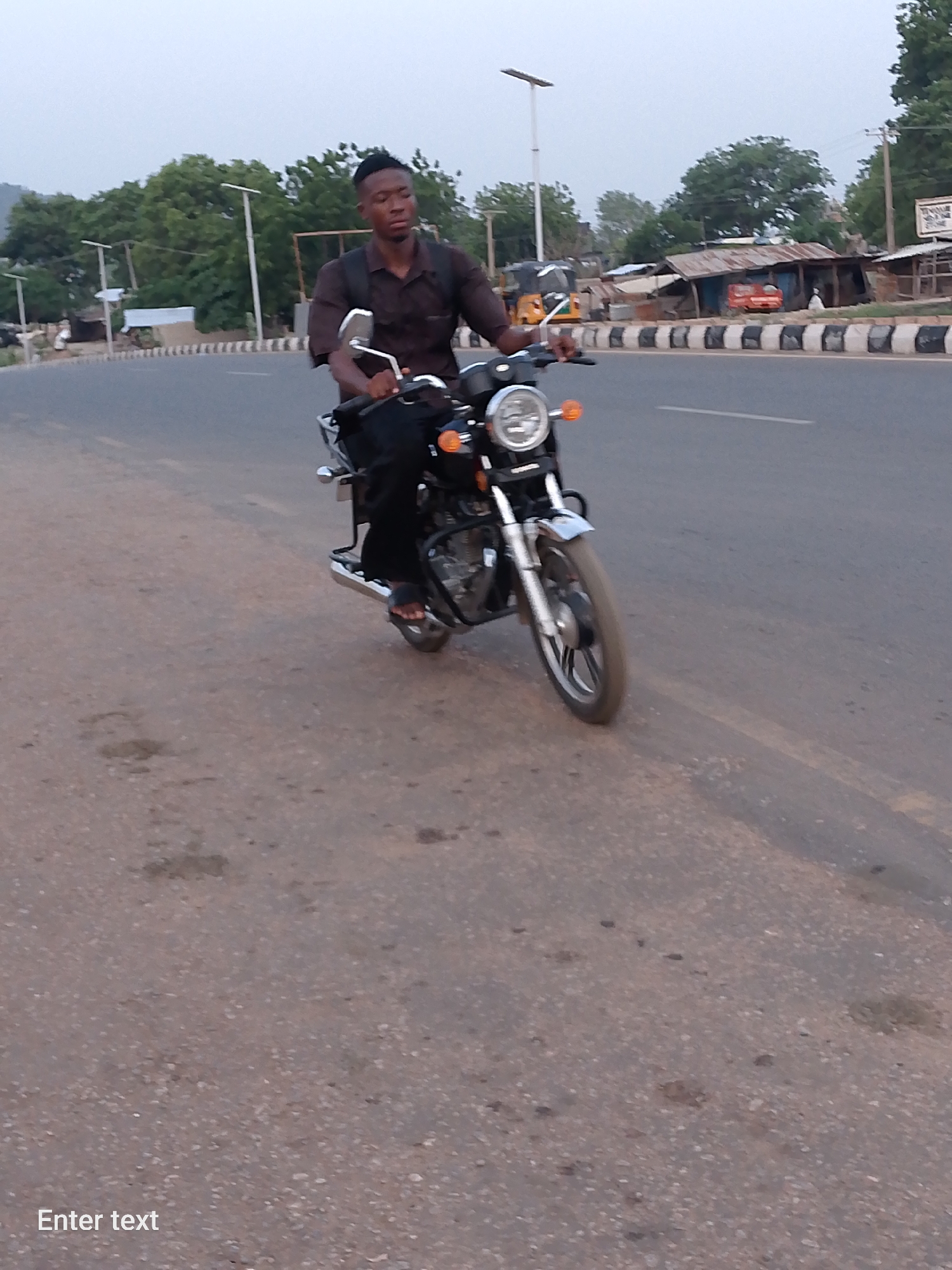 A man rides a black motorcycle along a paved road in Adankolo, Nigeria. He is wearing a brown short-sleeved shirt, black pants, and sandals, with a backpack. The road is bordered by a black and white striped curb. Buildings and trees are visible in the background under a grey sky.