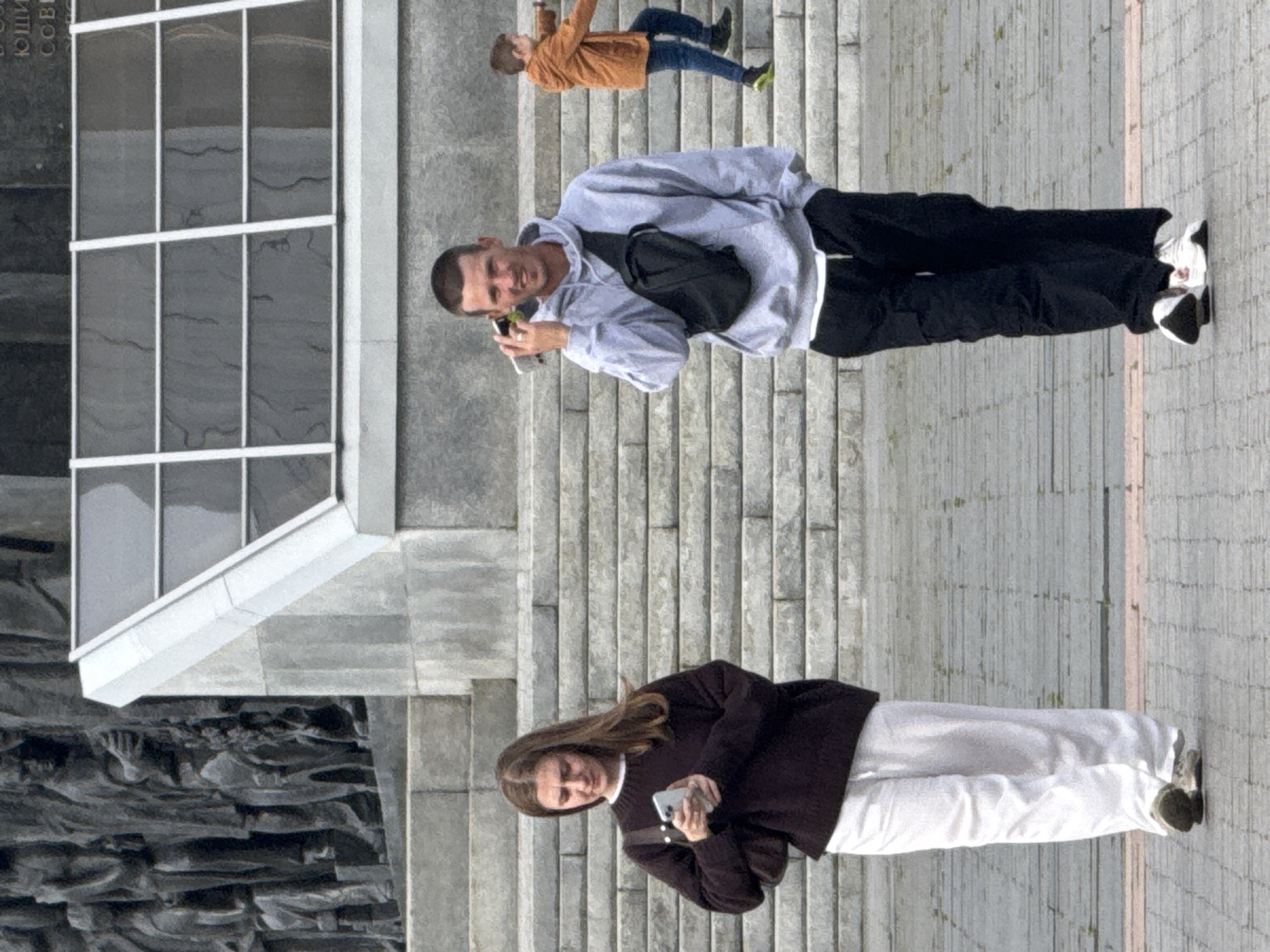 The image depicts an outdoor scene with several individuals on a paved area with large stone steps in the background. In the foreground, a young woman with long brown hair is walking to the left, looking down at her mobile phone. She is wearing a dark brown knitted sweater and white pants, carrying a dark brown shoulder bag. To her right, a man with short brown hair is walking towards the viewer, holding a mobile phone to his ear. He is wearing a light gray hooded sweatshirt, black cargo pants, and a black sling bag across his chest. In the background, a child wearing an orange jacket and blue jeans is ascending the stone steps to the right. The architecture in the background suggests a monumental or public space, possibly a memorial or government building. The overall atmosphere is casual and urban. The inscription on the monument in the background, though partially obscured, contains Cyrillic letters, suggesting the location is within a Russian-speaking country, and specifically Moscow, Russia, given the context of such public monuments.