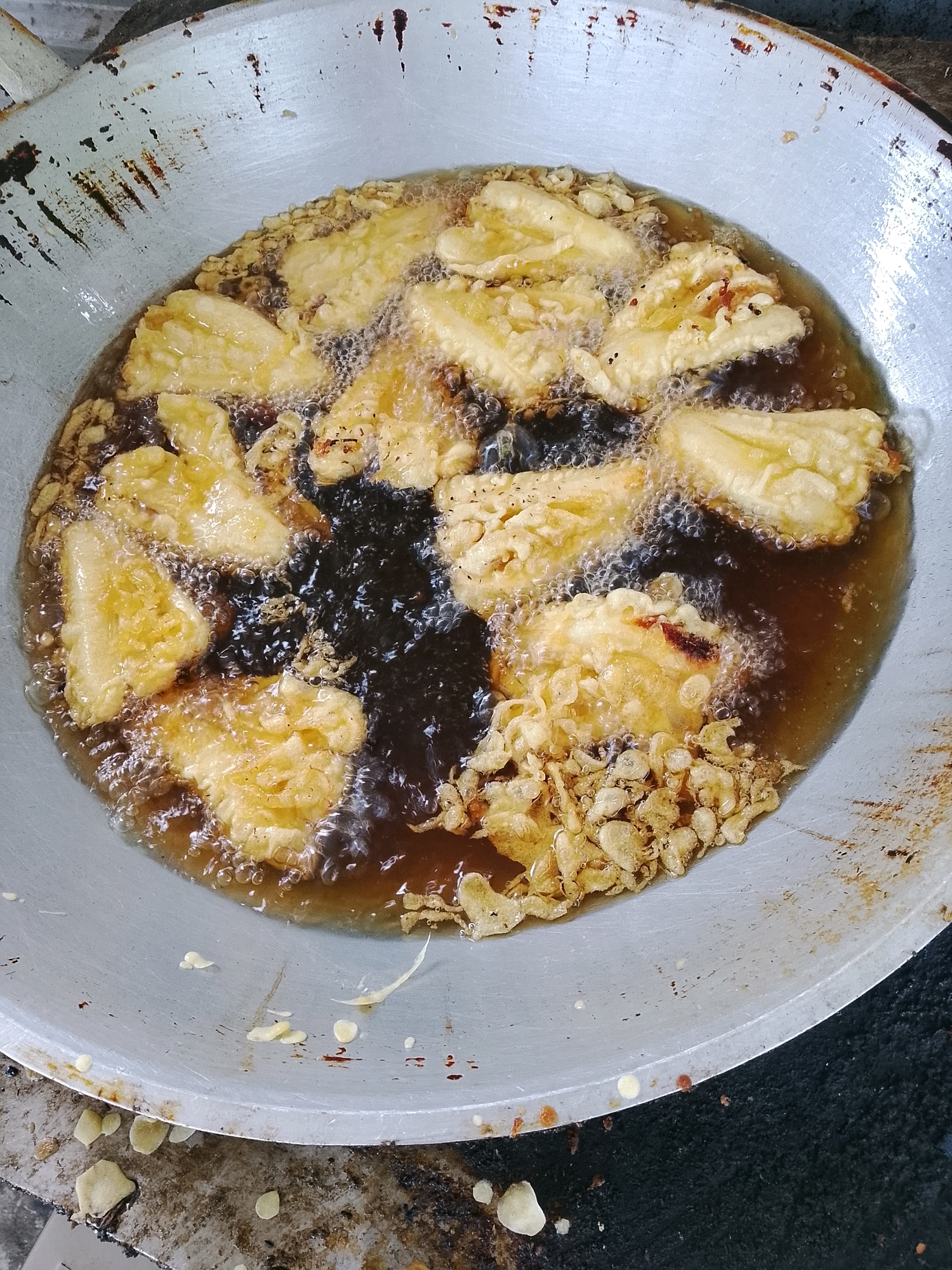In Banaran, Indonesia, a large, weathered metal wok is filled with hot oil, in which several pieces of battered food are being deep-fried. The food appears to be light yellow in color and some pieces are irregularly shaped, suggesting they may be bananas or a similar fruit. The oil is bubbling vigorously around the food, indicating a high temperature. Small pieces of the batter and possibly some other ingredients are floating in the oil, and some have adhered to the sides of the wok, which also shows signs of use and rust. The wok is resting on a dark, textured surface, possibly a stovetop or a griddle. The overall scene depicts a moment of food preparation, likely for a local dish given the context of Banaran.