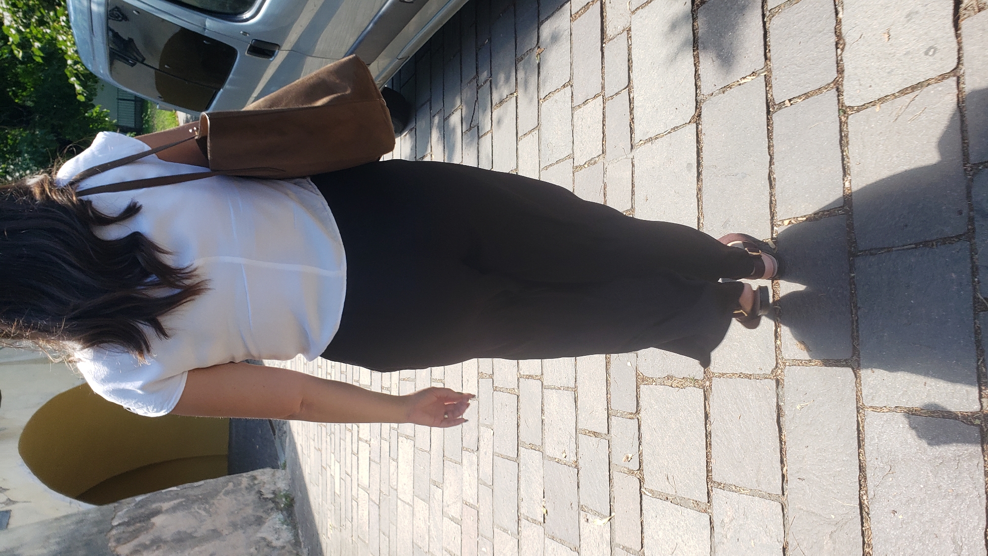A woman is walking away from the camera on a stone-paved street in Prešov, Slovakia. She is wearing a white short-sleeved blouse and black wide-leg pants. A brown tote bag is slung over her right shoulder. Her left hand is visible, with a ring on her finger. The street is lined with large, rectangular stones. To her left, there is an arched entryway to a building. To her right, a silver van is parked. Sunlight casts shadows on the ground.