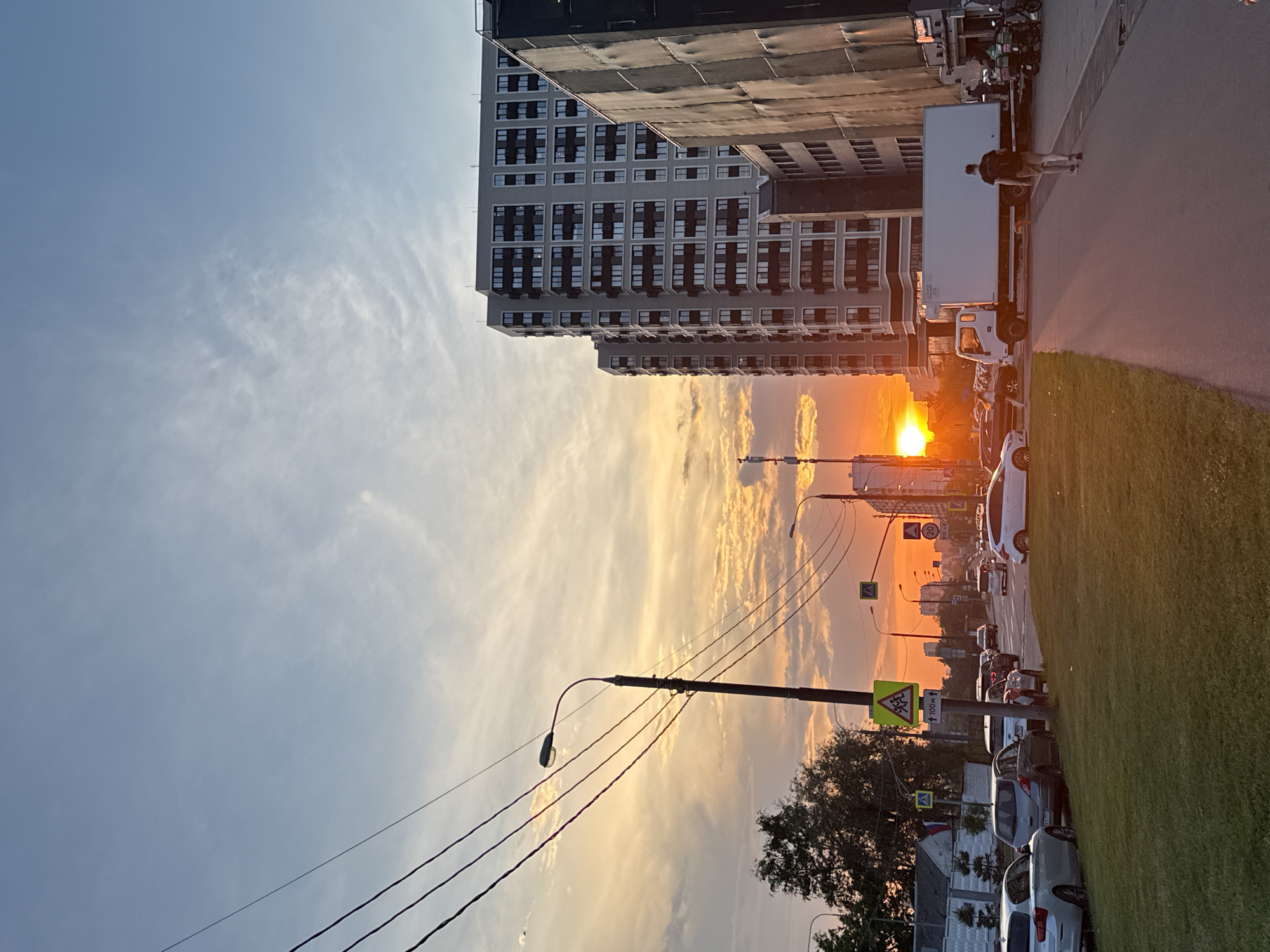The image depicts a sunset scene in a Moscow, Russia urban environment, likely a residential or mixed-use district. The sky is a gradient of blue, transitioning to orange and yellow hues near the horizon where the sun is setting, partially obscured by clouds. Tall, modern apartment buildings with numerous windows are visible on the right side of the frame. A white delivery truck is parked on the right side of the road, and a man with a backpack is walking away from the camera on the sidewalk. Several cars are parked along the street and on the grassy median. Traffic signals and streetlights are present. Power lines stretch across the upper left portion of the image.