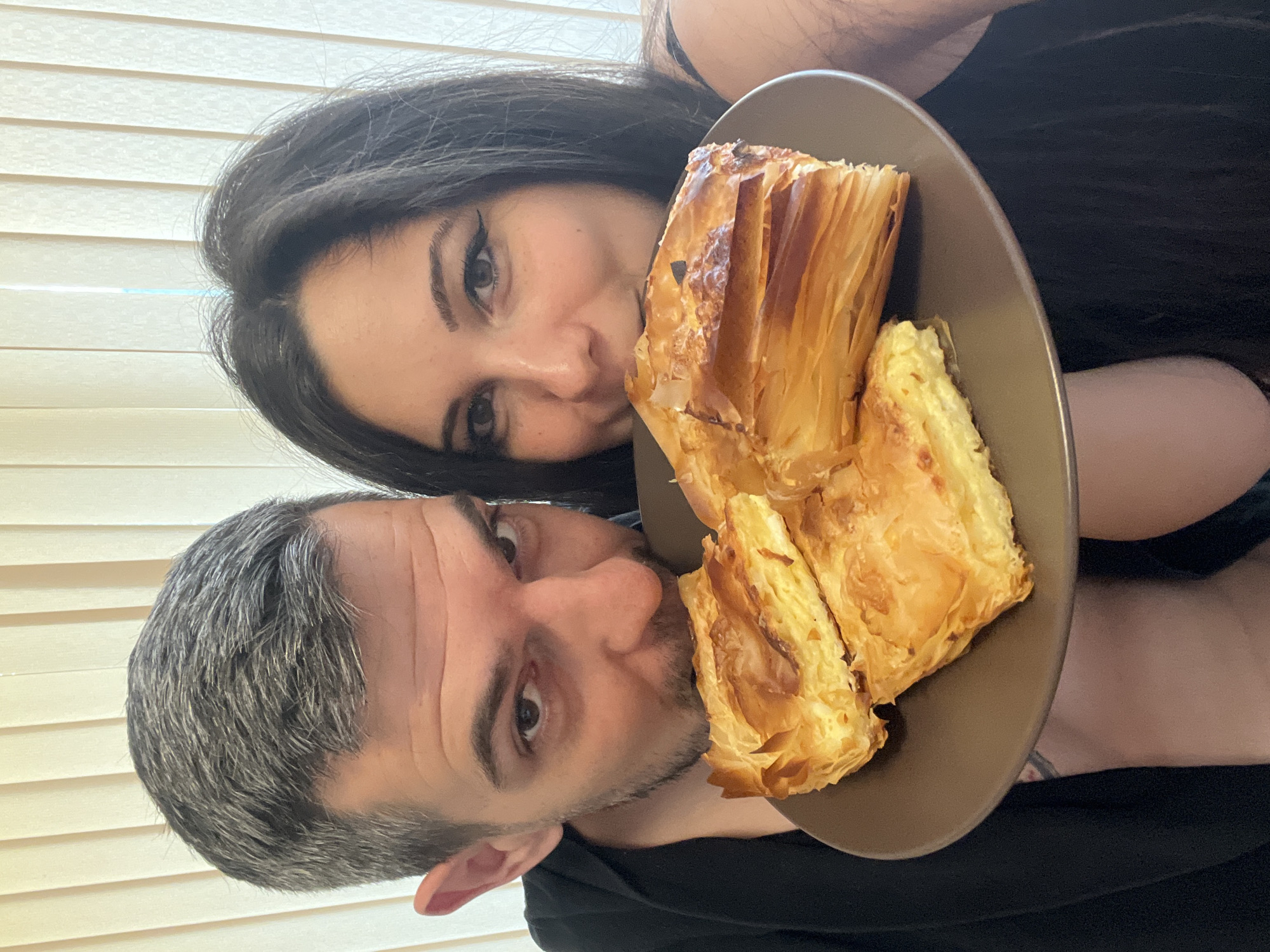 Two individuals, a male and a female, are shown in a close-up shot holding a plate with several pieces of baked goods. The male, on the left, has short, grey-streaked dark hair and is wearing a black top. He is looking towards the camera with a slight smirk. The female, on the right, has long, dark hair and is also wearing a dark top. She is smiling and looking towards the camera. The baked goods appear to be a type of pastry with flaky layers and a creamy filling, possibly a burek or spanakopita. The plate is a circular, matte brown color. The background consists of vertical blinds, suggesting an indoor setting. The lighting is soft and diffused. The image appears to have been taken in a residential setting in Sofia, Bulgaria, given the typical interior design elements.