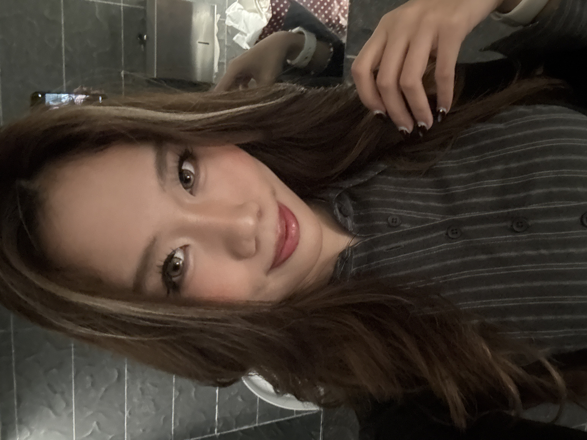 A young woman is posing indoors in Jiangbeicheng Subdistrict, China. She has brown hair with blonde highlights and is wearing a grey and white striped button-down shirt. Her nails have black and white tips. Her makeup includes pink blush and lipstick. A hand with painted nails touches her hair. A cell phone is visible in the background.