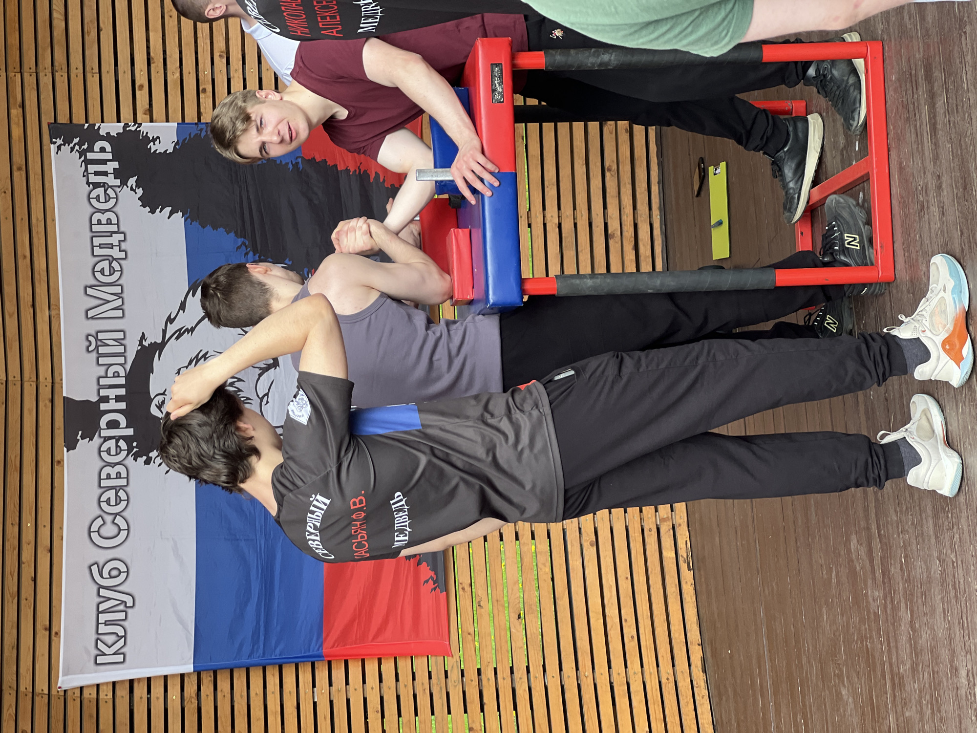 The image shows a scene from what appears to be an arm wrestling competition. Two young men are arm wrestling at a table with a blue and red pad. A man stands nearby observing the match. The backdrop features a flag in the colors of the Russian flag, along with the name "Клуб Северный Медведь", meaning "Northern Bear Club" in Russian. The location is Moscow, Russia. The ground is wooden planks, indicating an outdoor or covered area, such as a patio or porch.