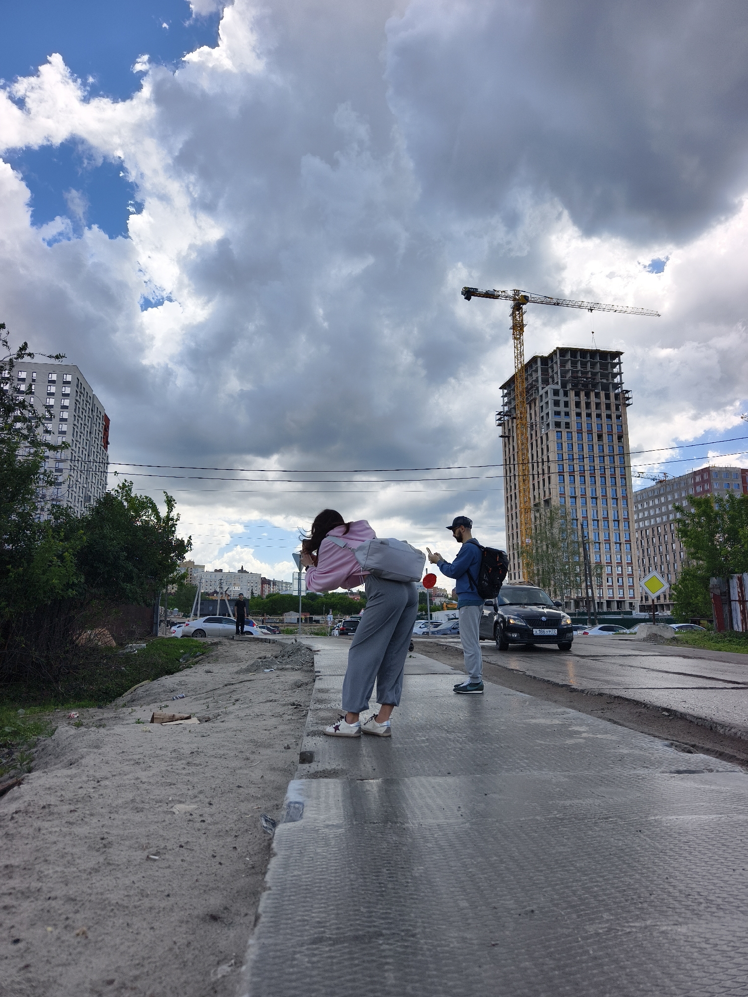 A wide, ground-level shot captures a cityscape under a sky dominated by a mix of blue patches and heavy, gray clouds. In the foreground, a woman wearing a light pink top, gray pants, and white sneakers is bent over, holding a phone up to take a photo. Beside her and slightly behind stands a man wearing a blue shirt, gray pants, sneakers, a face mask, and a baseball cap, with a backpack slung over his shoulder.
The pair stands on what appears to be a temporary or makeshift platform laid across a gap in the pavement, its surface appearing slightly wet. Beyond the platform is a busy road with cars passing by.
In the background, a tall, modern building under construction is visible. A crane towers above it. Surrounding the construction site are other residential buildings, trees, and various urban elements.
The license plate of the black car driving by shows "X 186 YP 72" indicates a vehicle registration in Tyumen, Russia. The photo was likely taken in Tyumen.