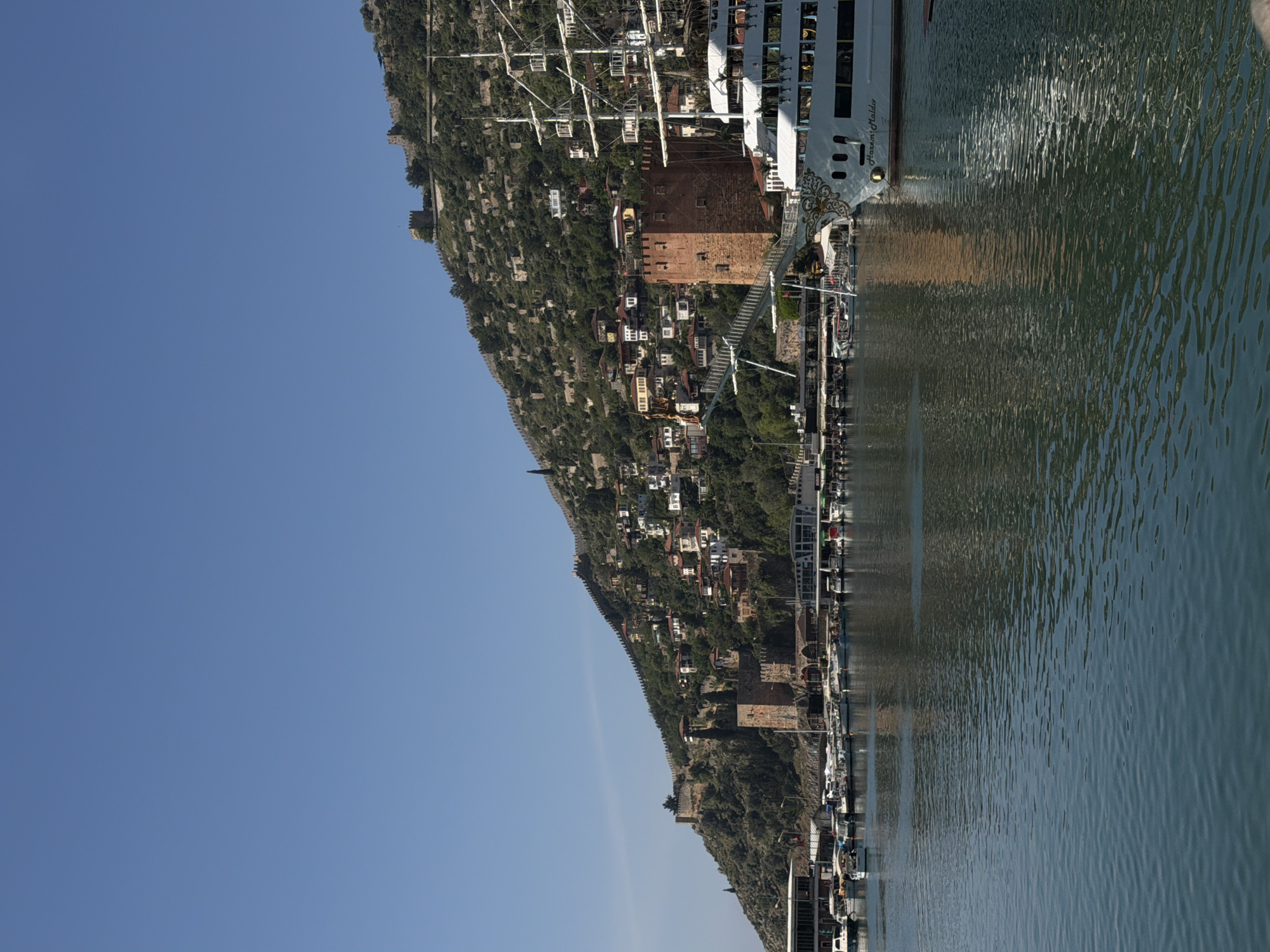 The image shows Alanya Castle and the Red Tower in Alanya, Turkey, from a boat. The medieval castle is built atop a rocky peninsula, with defensive walls snaking down the hillside. The Red Tower, a prominent red-brick octagonal structure, stands near the harbor. The clear blue sky contrasts with the green hillside dotted with buildings and trees. A cruise boat is docked along the waterfront, its reflection visible in the water.