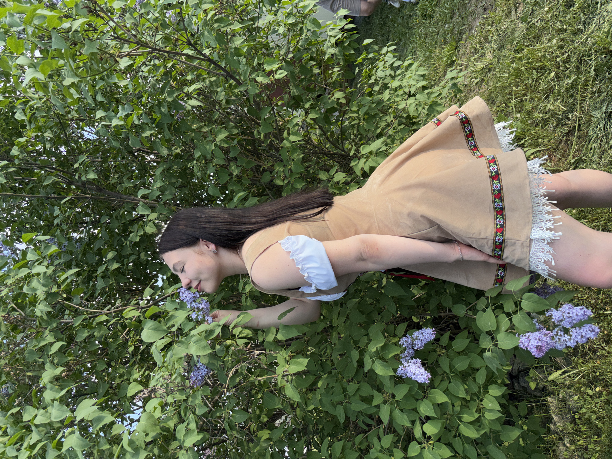 A woman in a tan dress with white lace trim and a floral ribbon design is smelling purple lilac flowers on a bush. The photo appears to be taken in an outdoor, grassy area. The city is Пальники, Russia.