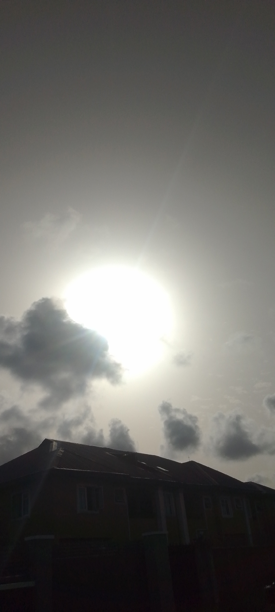 The image shows a two-story building silhouetted against a bright, overcast sky. Clouds surround a bright sun, obscuring direct sunlight. The building's roofline and windows are visible, but details are obscured by the darkness. The location is in the city of Unknown, Unknown.